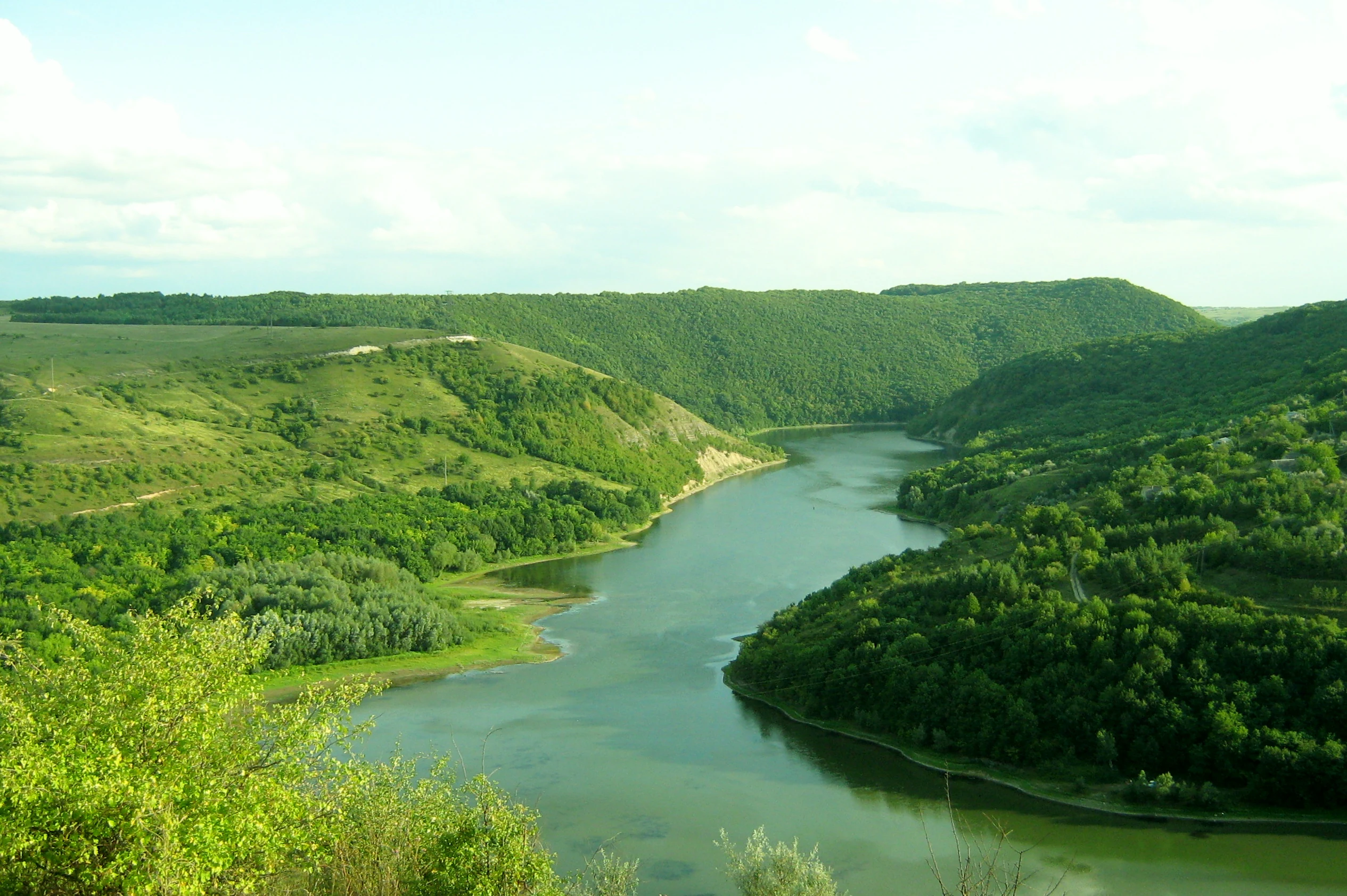 A wide river flowing through a valley with green hills and trees on both sides under a partly cloudy sky