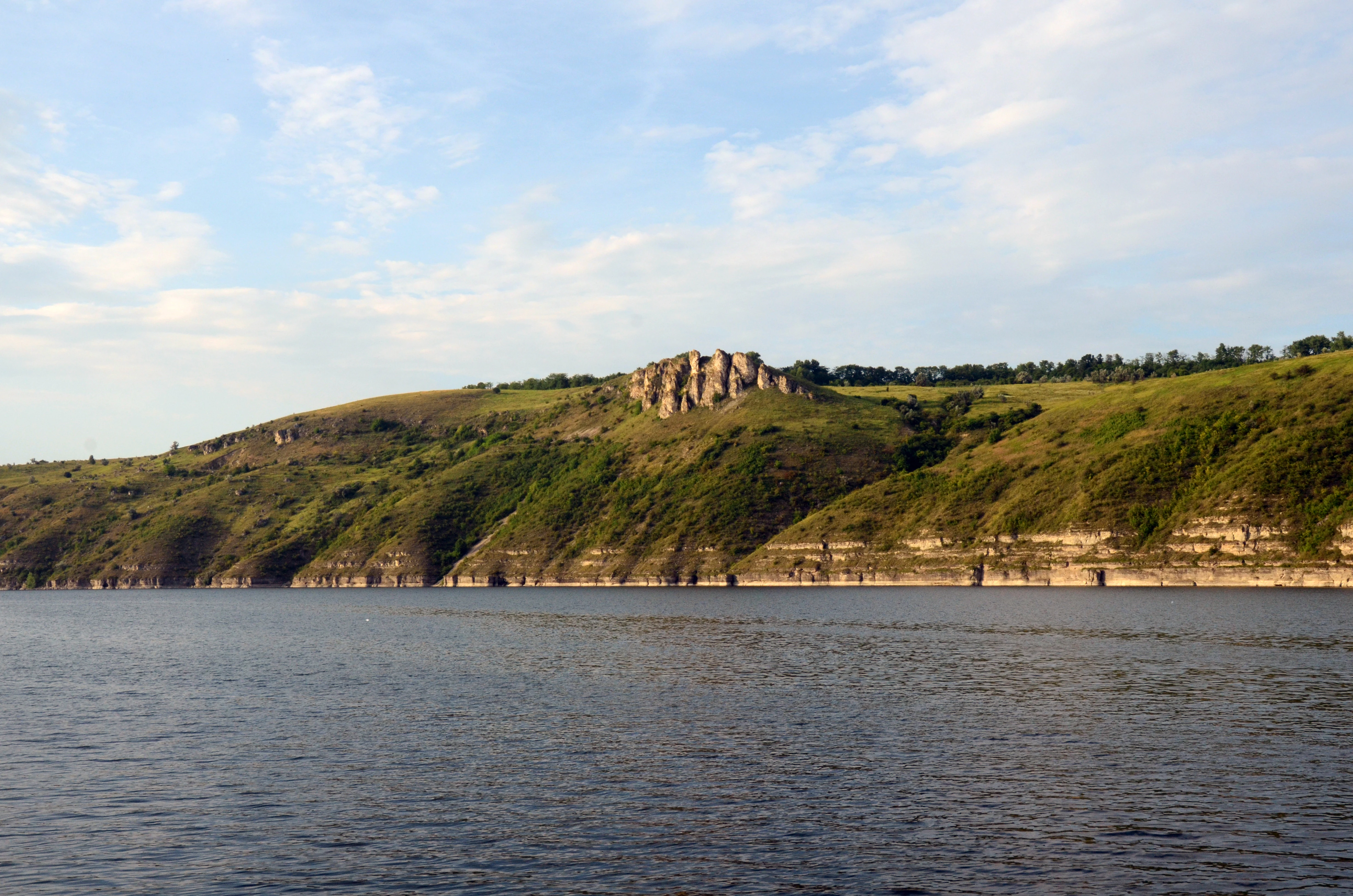 Aerial view of a wide river with rolling green hills and rocky cliffs in the background under a partly cloudy sky