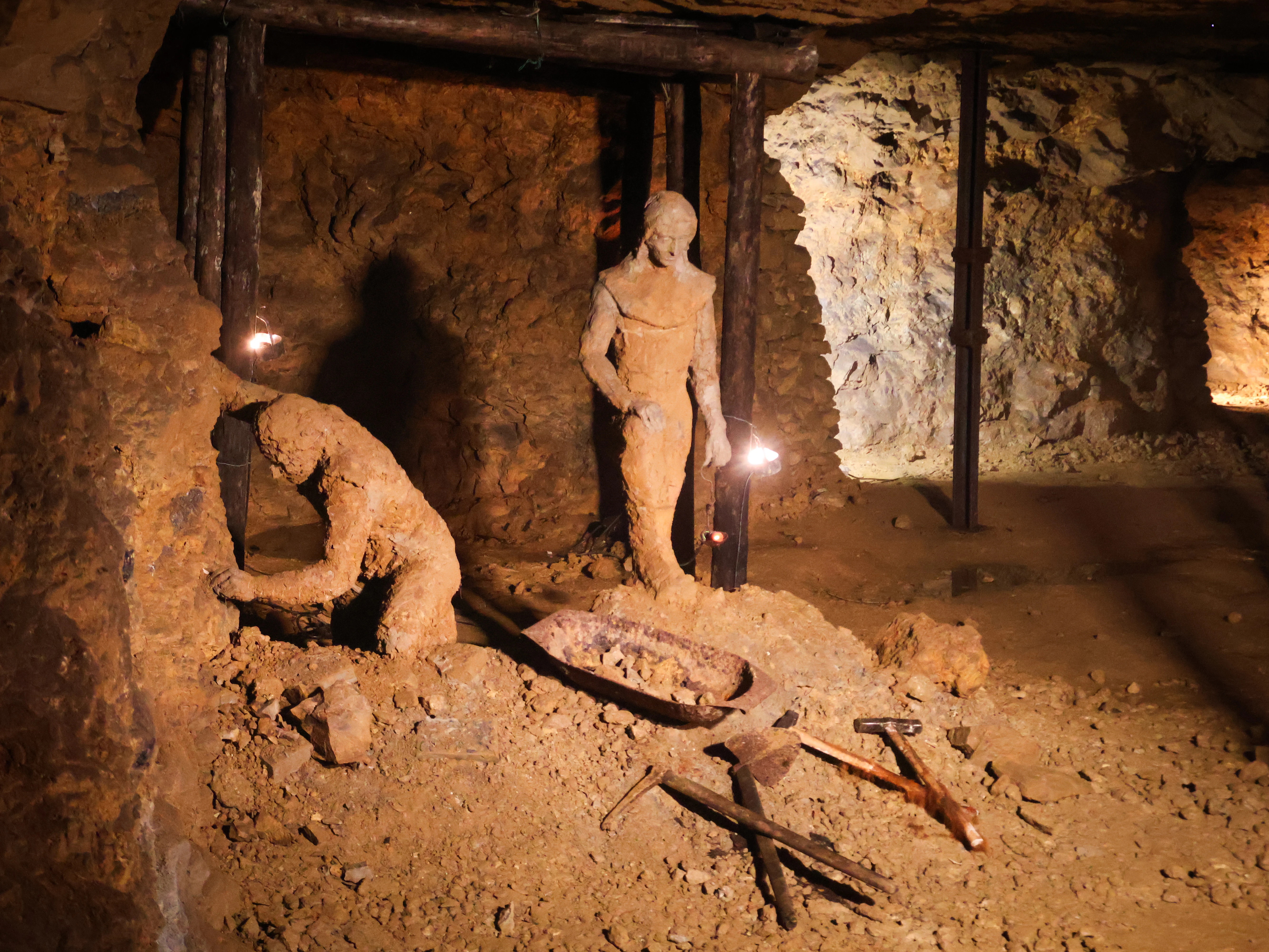 A dimly lit underground mine tunnel featuring two life-sized clay-colored figures depicting miners at work; one figure is bent over working the rock wall, while the other stands nearby, with period tools and a cart in the foreground.