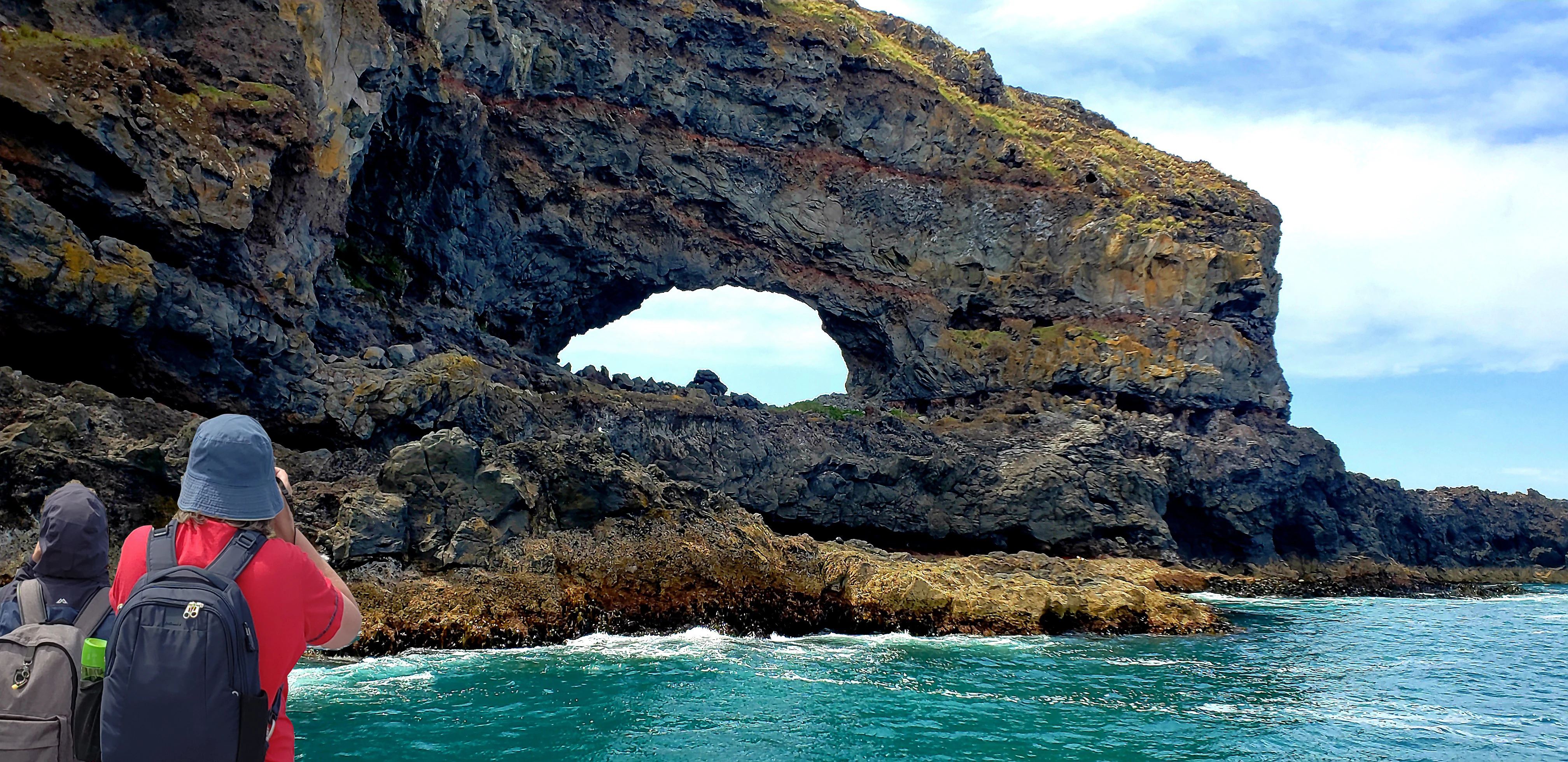 A view from the water showing a large natural stone archway in a coastal cliff face, with two people in the foreground on a boat looking toward it.