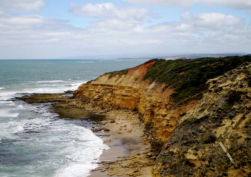 Acantilados de roca en capas con vegetación, con vistas a las olas del océano que rompen en una playa de arena bajo un cielo parcialmente nublado.