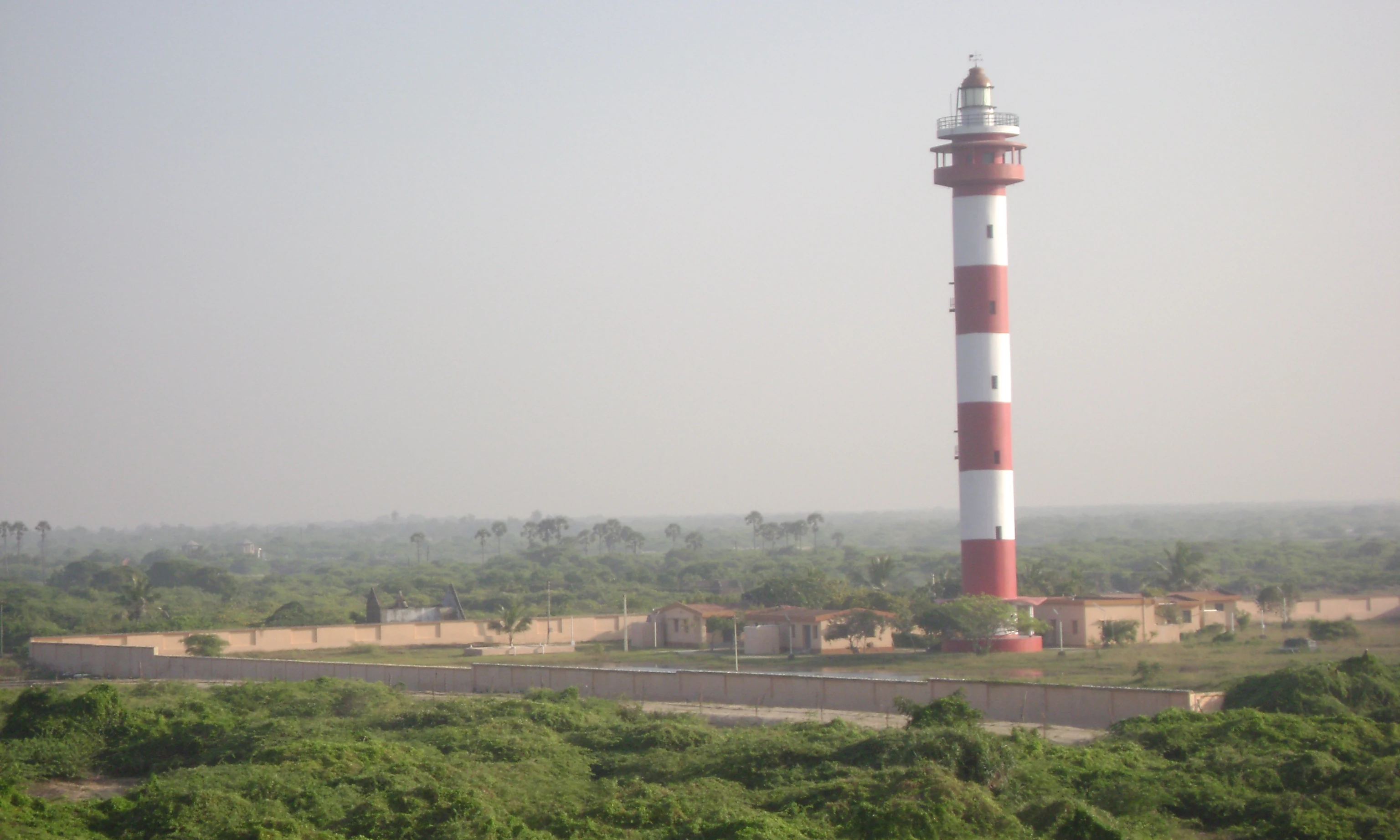 A tall, red and white striped lighthouse standing behind a perimeter wall in a coastal landscape with green vegetation under a hazy sky.