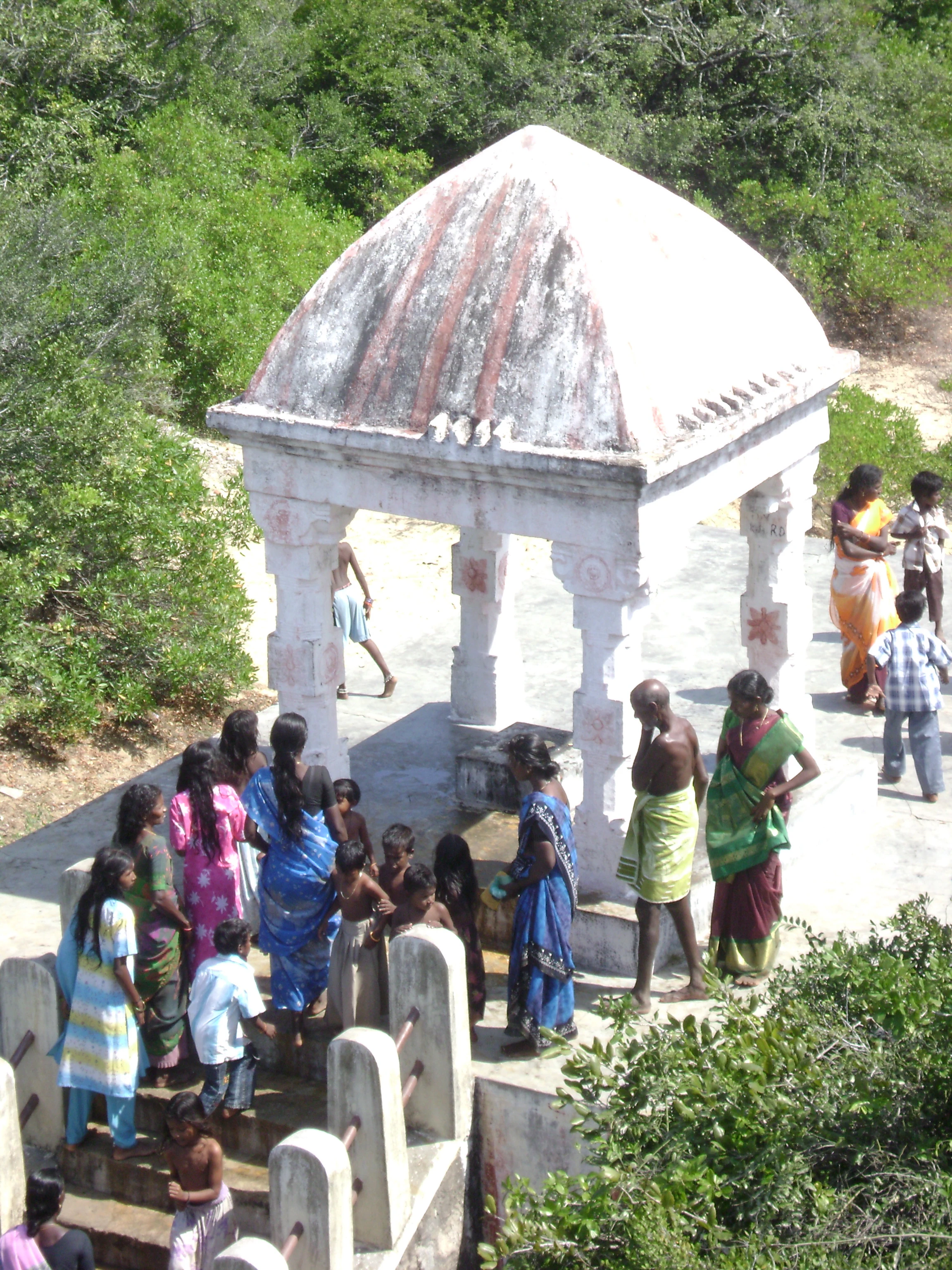 A group of people gathering at a small white stone shrine with a domed roof in a forested area.