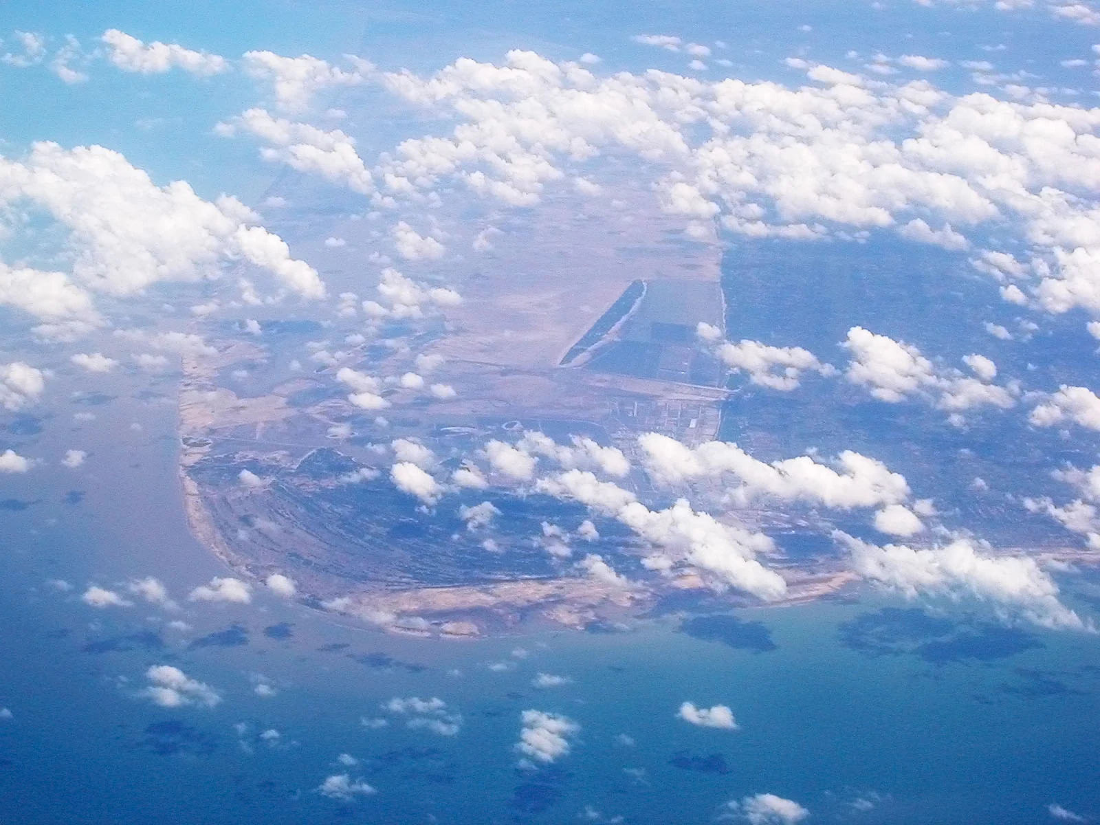An aerial view from an aircraft looking down at a coastal landscape with sandy shoreline, wetlands, and open water, partially obscured by scattered clouds.