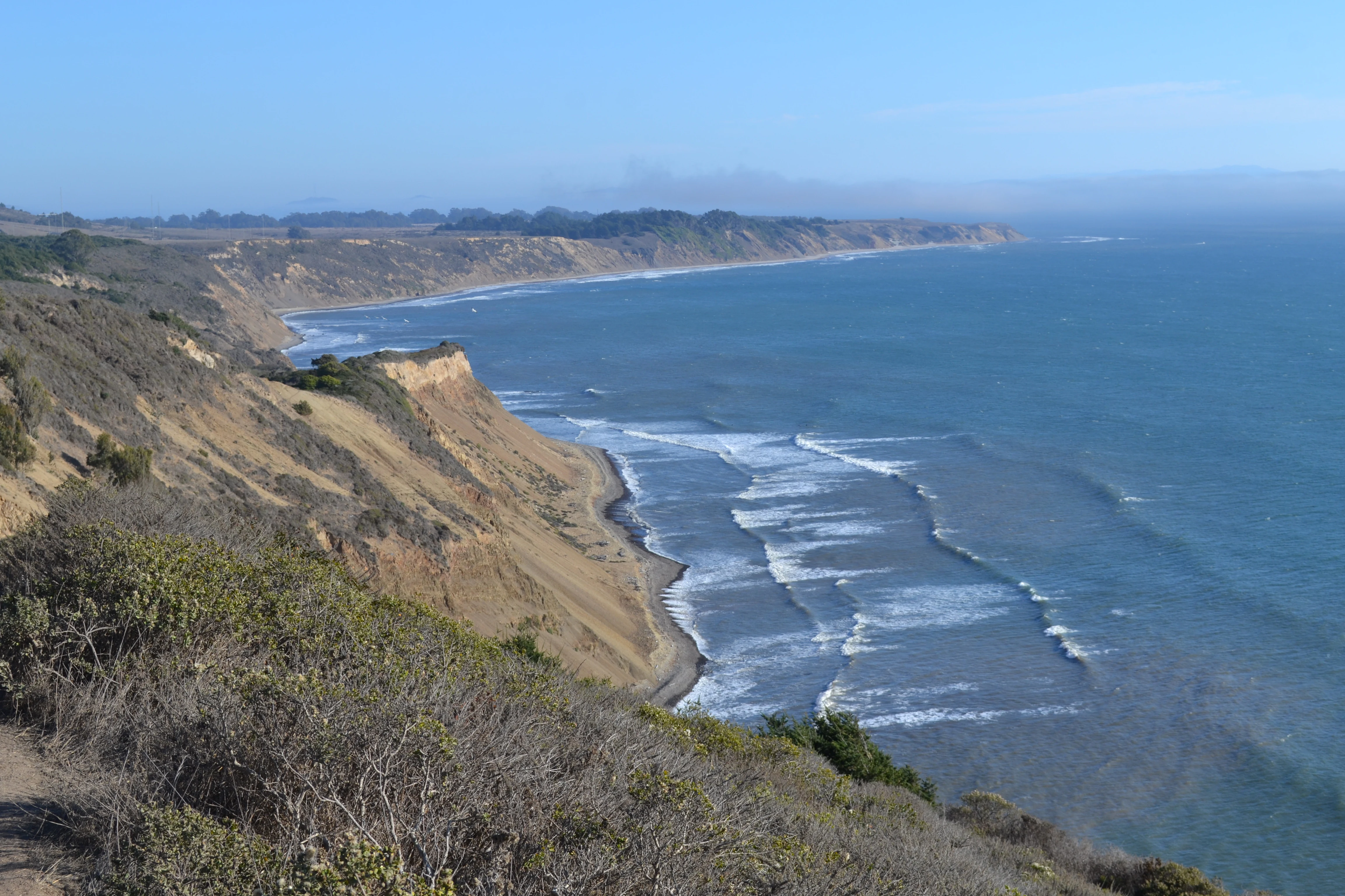 Coastal view from a high vantage point overlooking rugged cliffs, a narrow beach, and the Pacific Ocean.