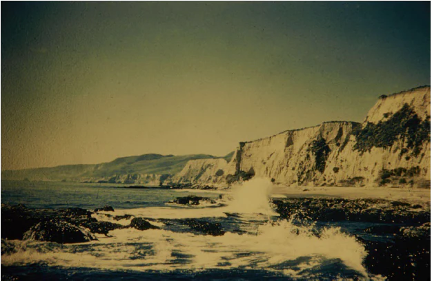 A vintage color photograph of the Point Reyes coastline showing steep, light colored cliffs meeting the ocean with waves crashing against rocks in the foreground.