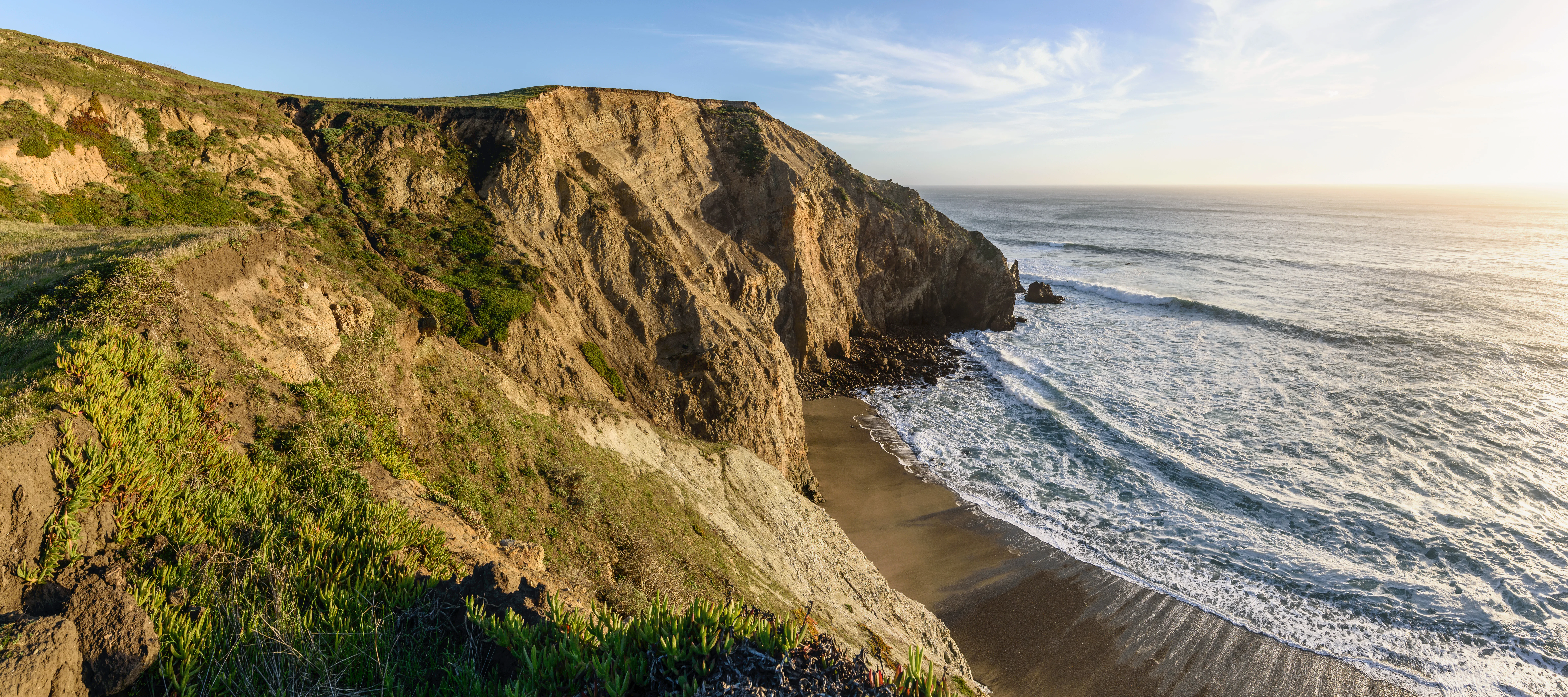 Panoramic view of rugged coastal cliffs descending into the Pacific Ocean, with crashing waves hitting a sandy beach under bright sunlight.