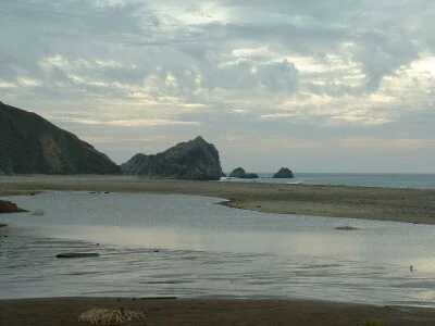 A wide, eye-level view of a sandy beach at low tide with a small, calm pool of water in the foreground, rugged cliffs on the left, and rocky sea stacks rising out of the ocean in the distance under a cloudy sky.