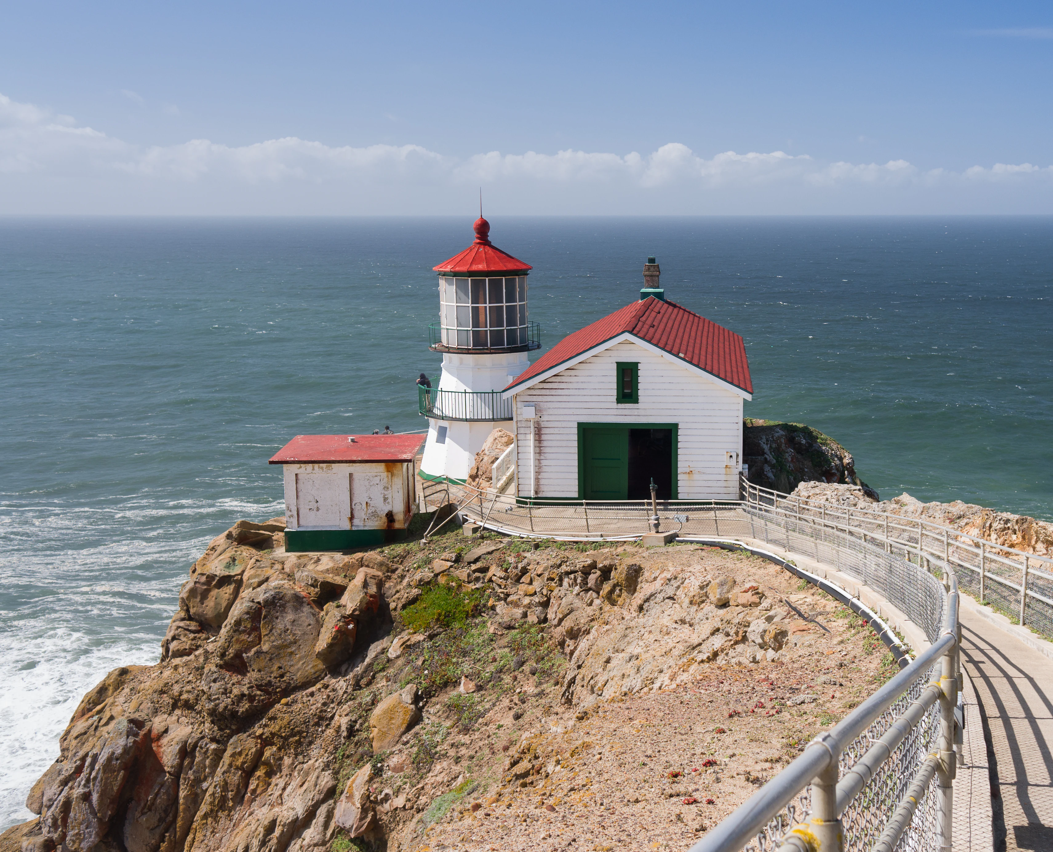 Point Reyes Lighthouse with a white exterior and red roof, situated on a rocky cliffside next to the ocean, with a metal railing along a walkway in the foreground.