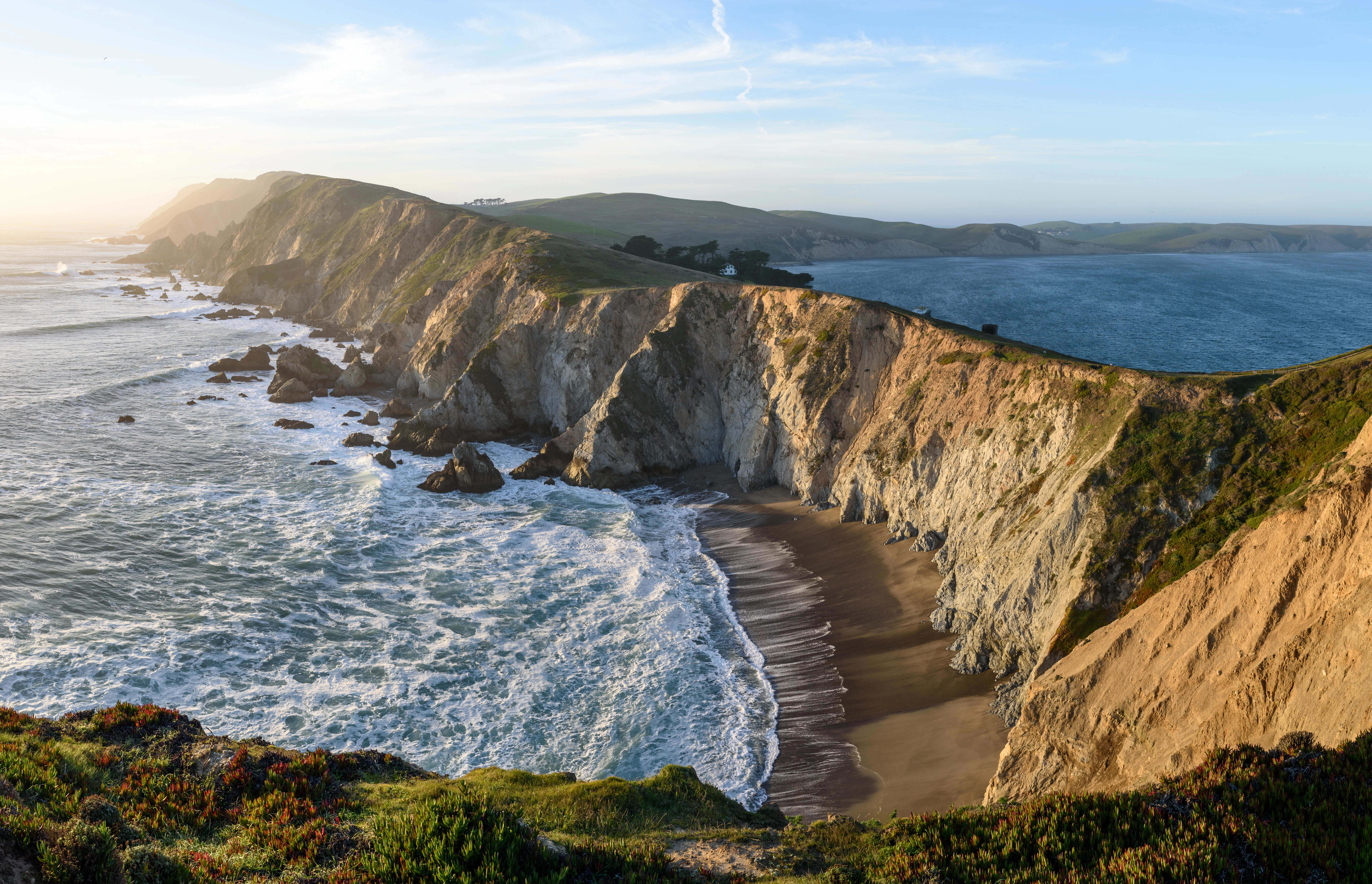 A wide panoramic shot of coastal cliffs meeting the Pacific Ocean with waves crashing against the sandy beach below under a clear sky.