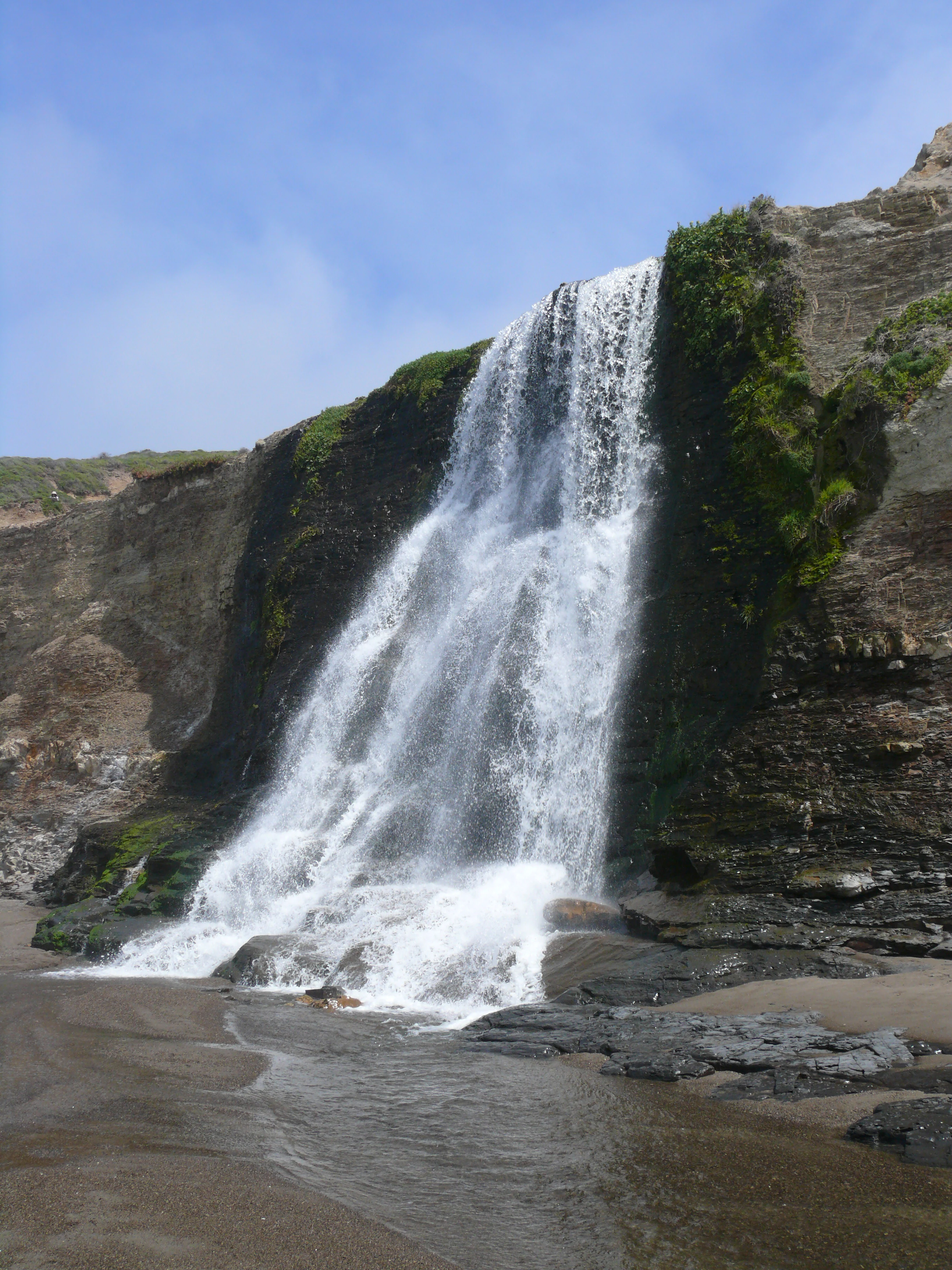 A waterfall cascading over rugged coastal cliffs directly onto a dark sand beach.
