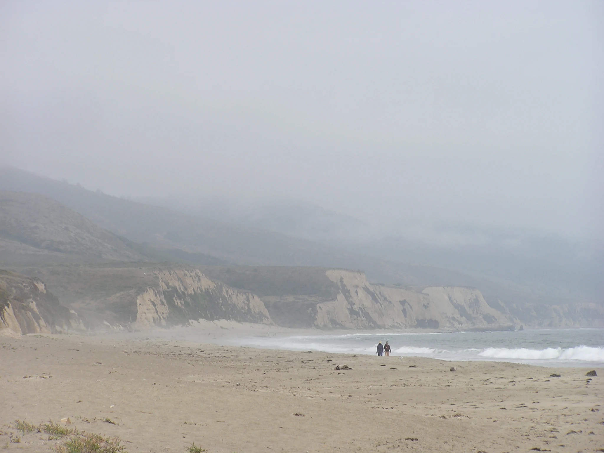 A wide, hazy view of a sandy beach bordered by steep coastal cliffs obscured by dense fog, with two distant figures walking near the surf.