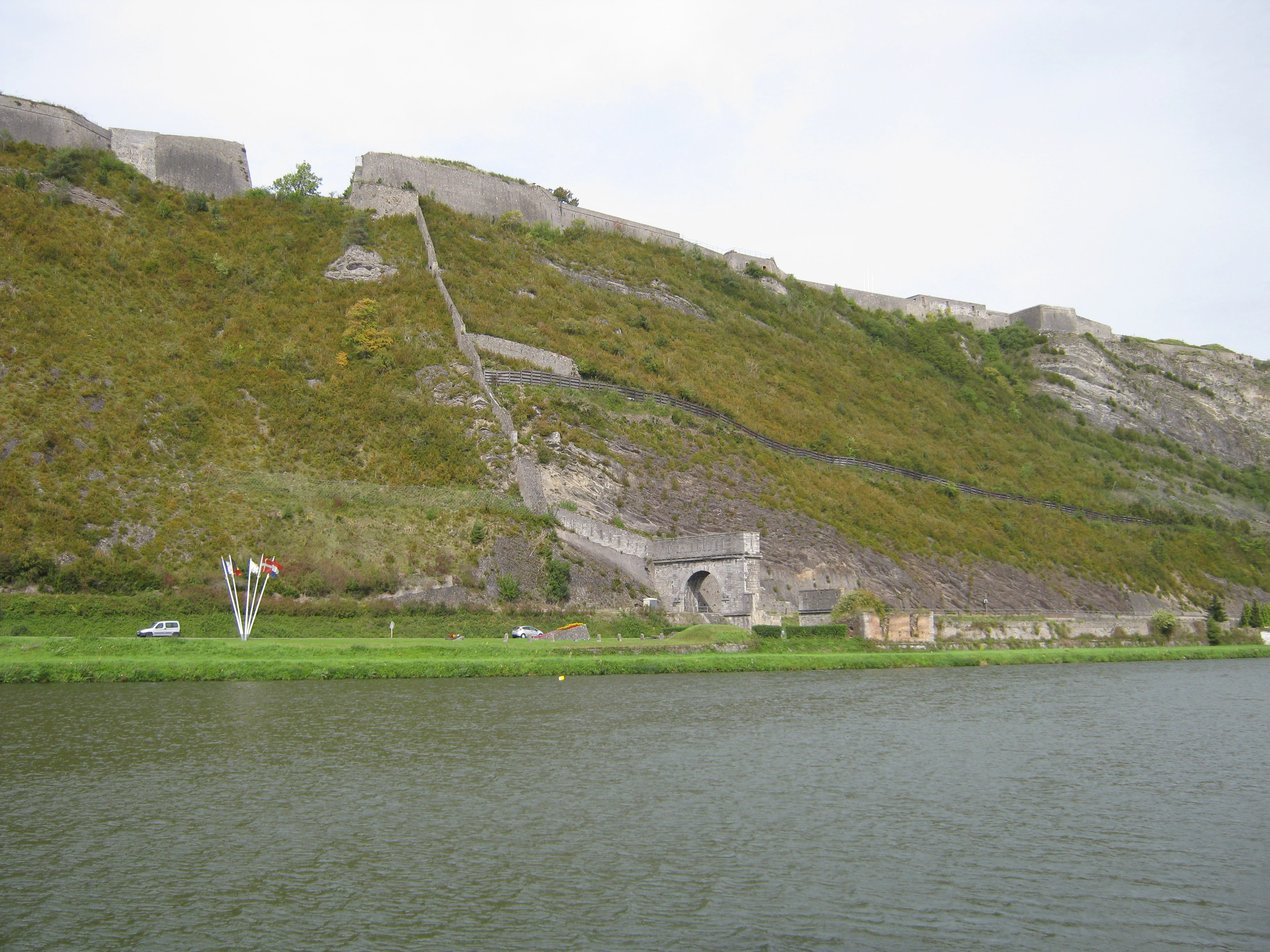 A green hillside rising from a riverbank, featuring a stone fortification wall and the Porte de France archway at its base.