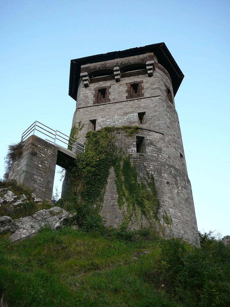 A historic stone watchtower built on a rocky cliffside with a small footbridge attached to one side and ivy growing on its walls.