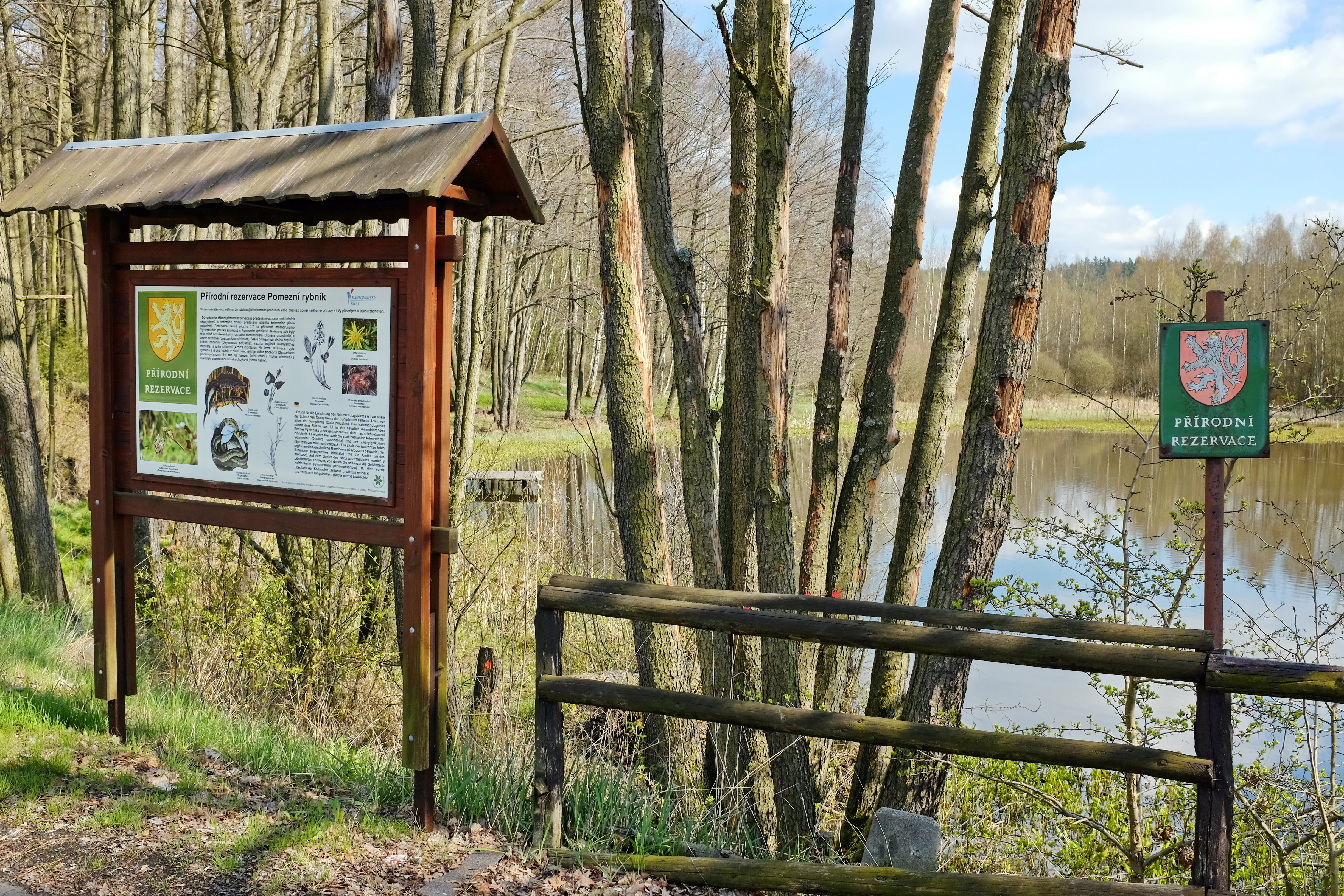 An informational wooden sign stand and a smaller green nature reserve sign stand positioned near the edge of a pond, surrounded by trees.