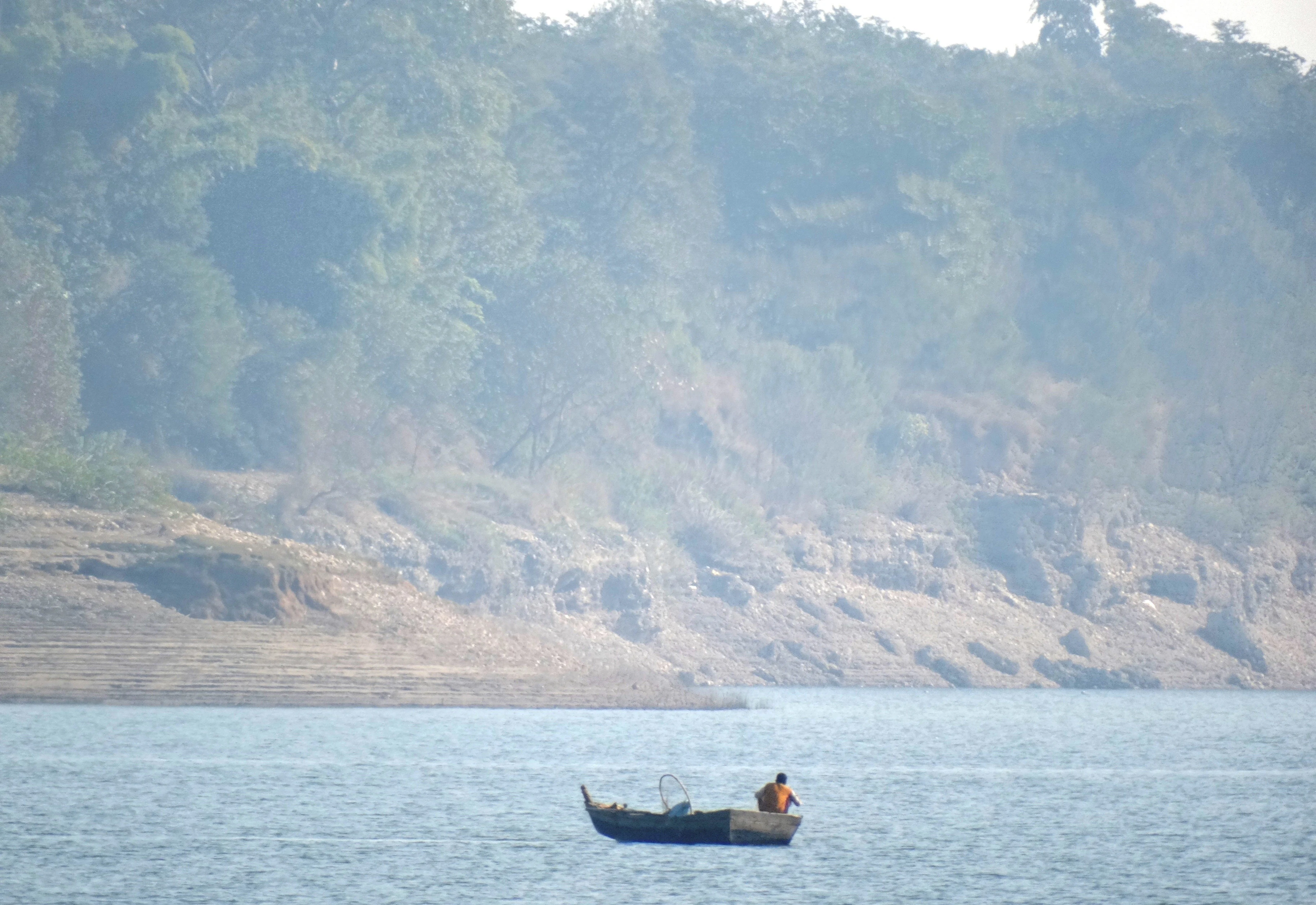A small boat with a lone fisherman on a calm lake with a forested hillside in the background.