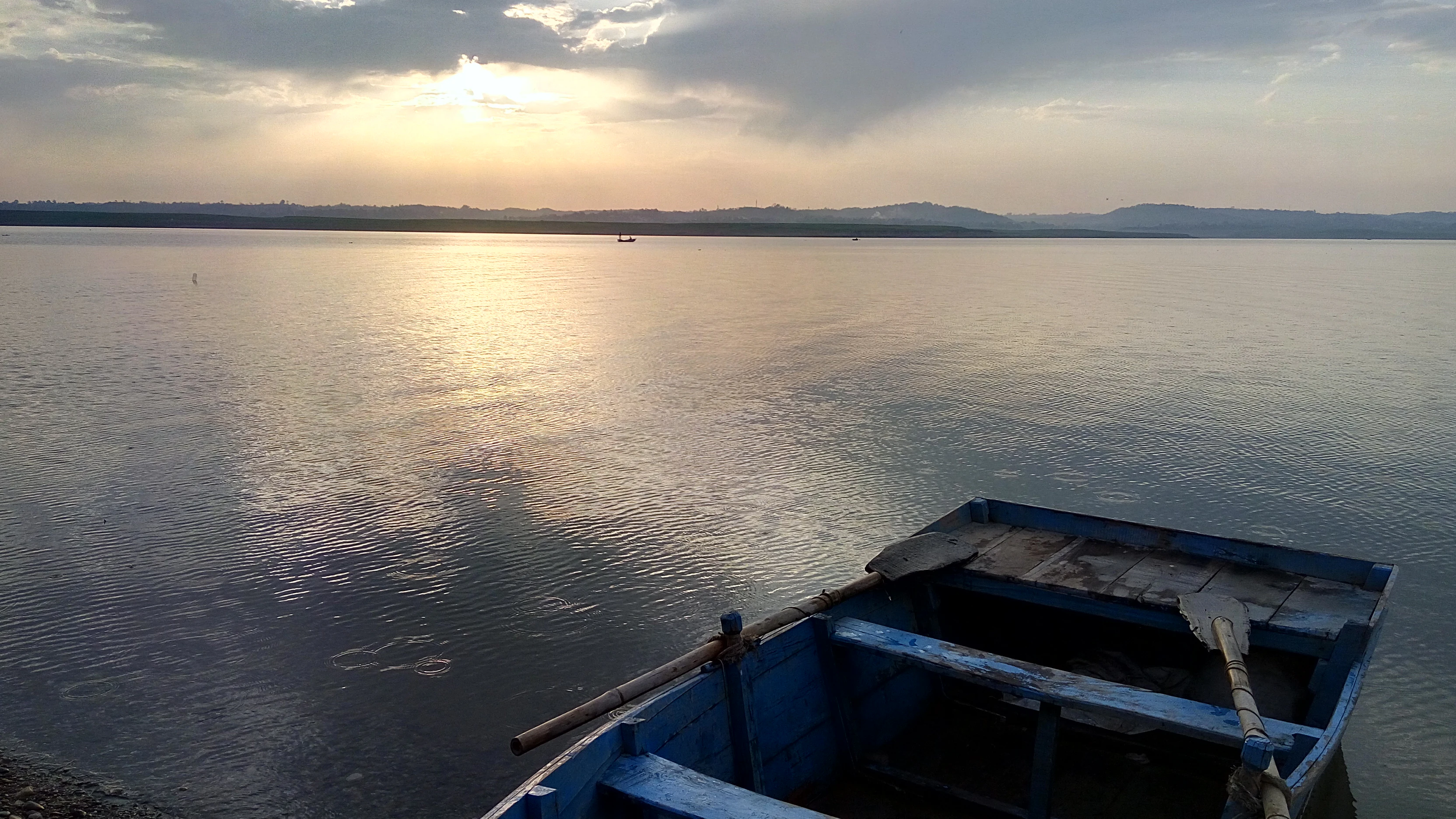 A small blue wooden rowboat sits in the foreground on a calm, expansive lake under a cloudy sunset sky, with distant hills visible on the horizon.