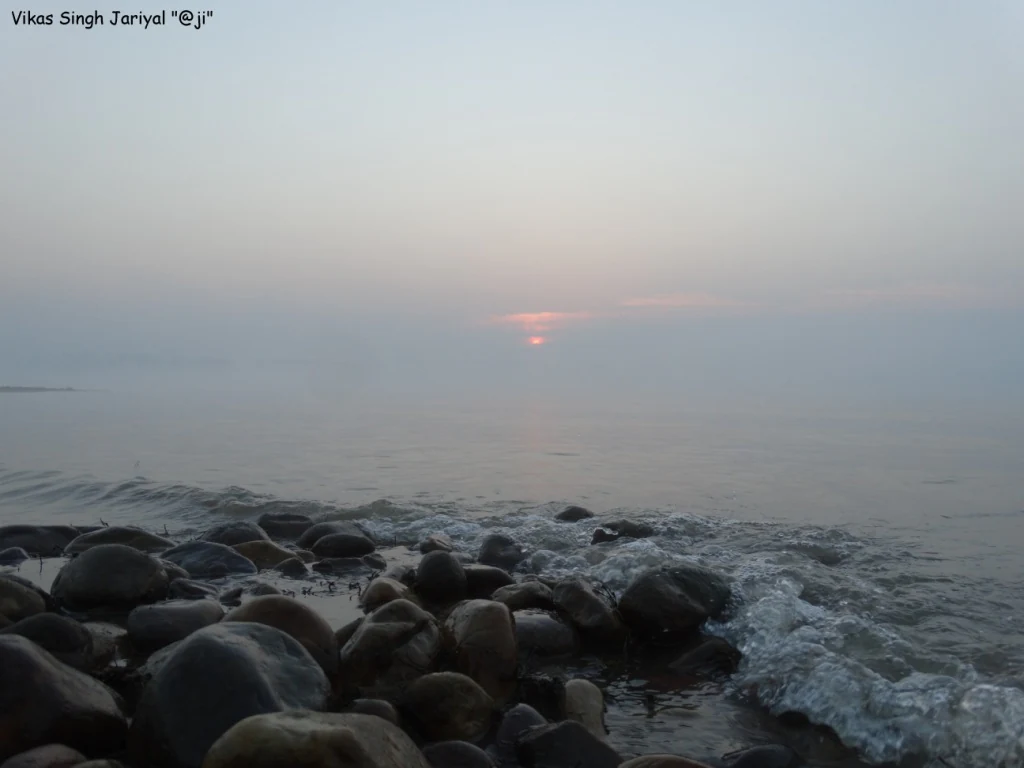 A wide, hazy sunrise view over the calm water of Pong Dam Lake, with smooth dark stones and gentle waves in the foreground.