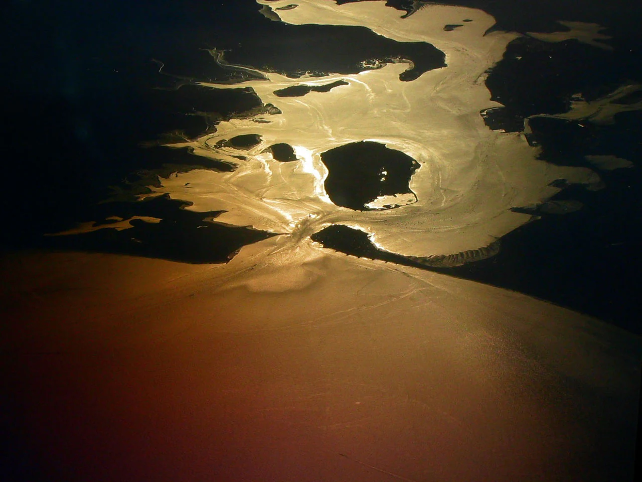 An aerial view of Poole Harbour at sunset, showing reflective waterways and several islands silhouetted against a golden, shimmering water surface.