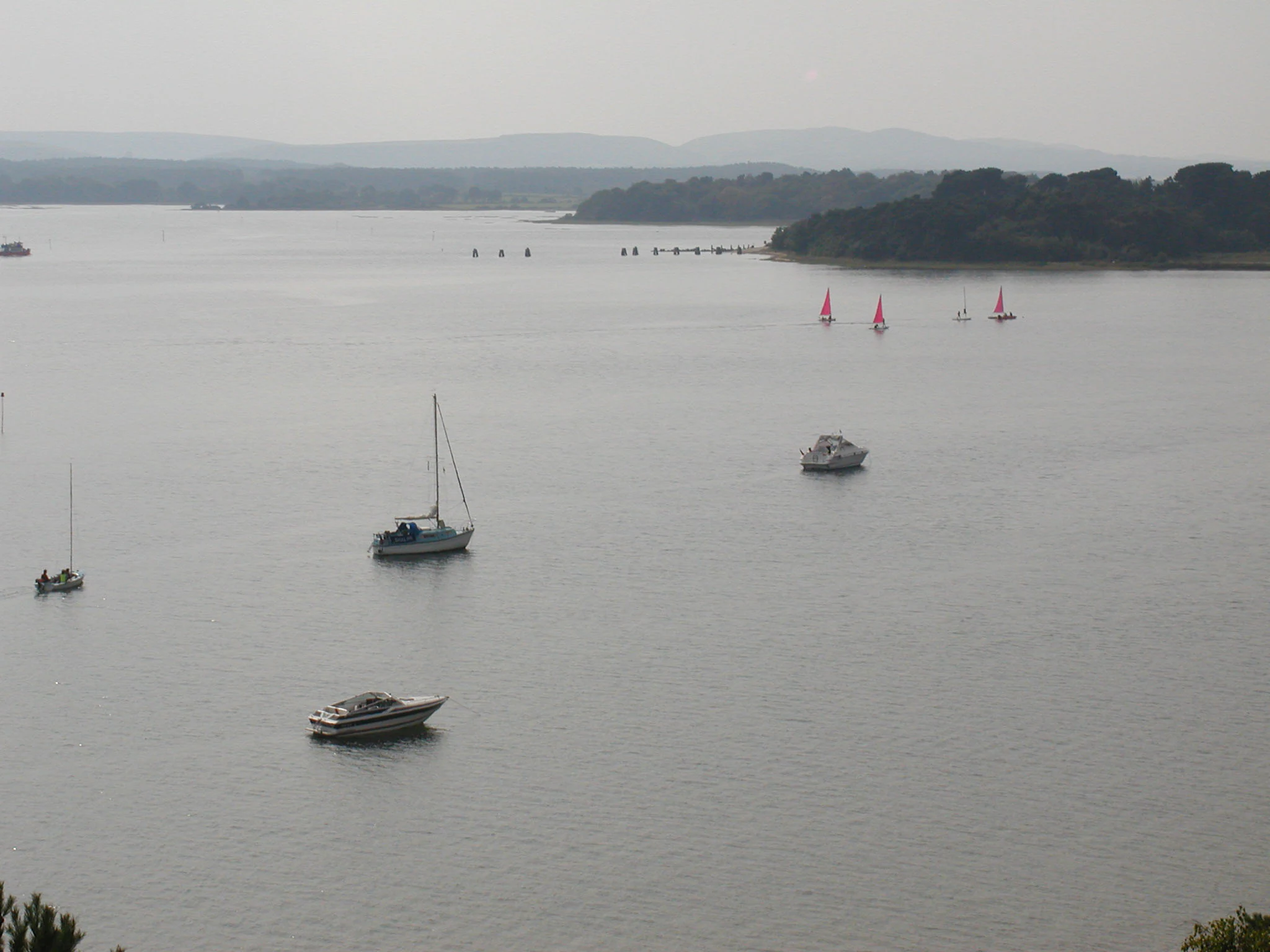 A hazy, elevated view across a large, calm harbour with several sailboats, motorboats, and small red-sailed vessels visible on the water.