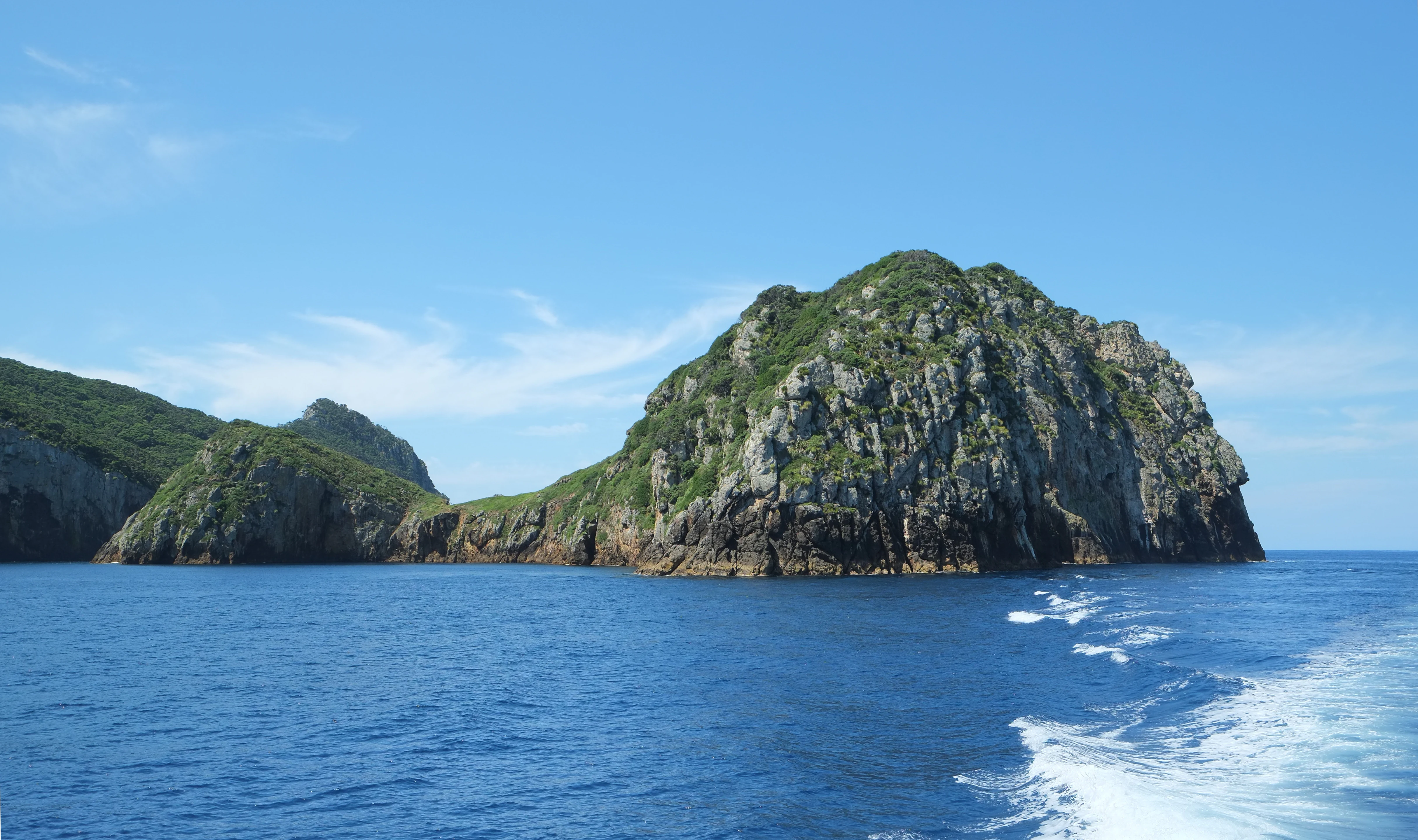 Islas rocosas escarpadas y cubiertas de vegetación emergen del profundo océano azul, vistas desde una embarcación en movimiento con una estela visible en primer plano.