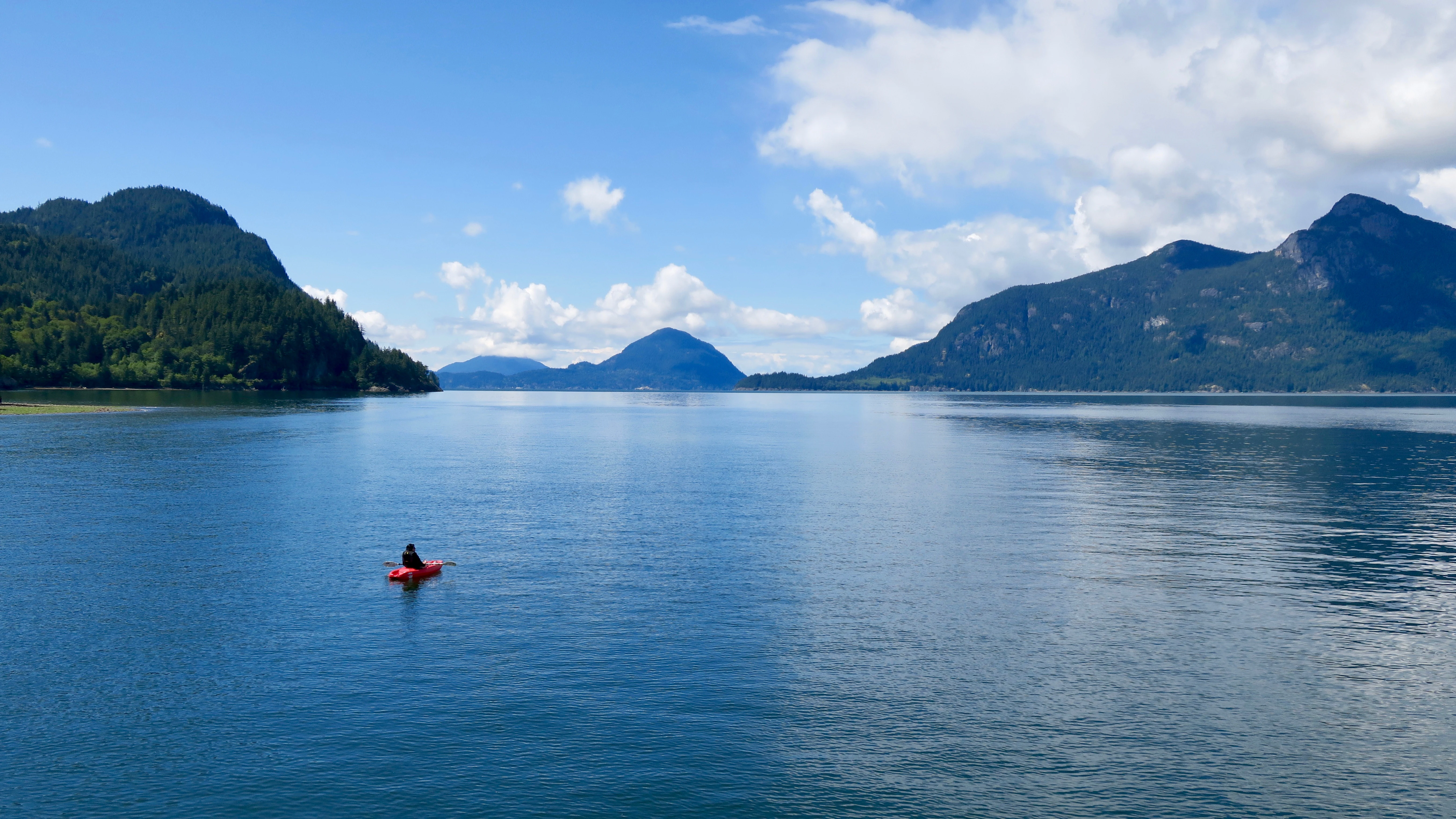 A wide angle view of a person in a red kayak on deep blue water, surrounded by forested mountains under a blue sky with white clouds.