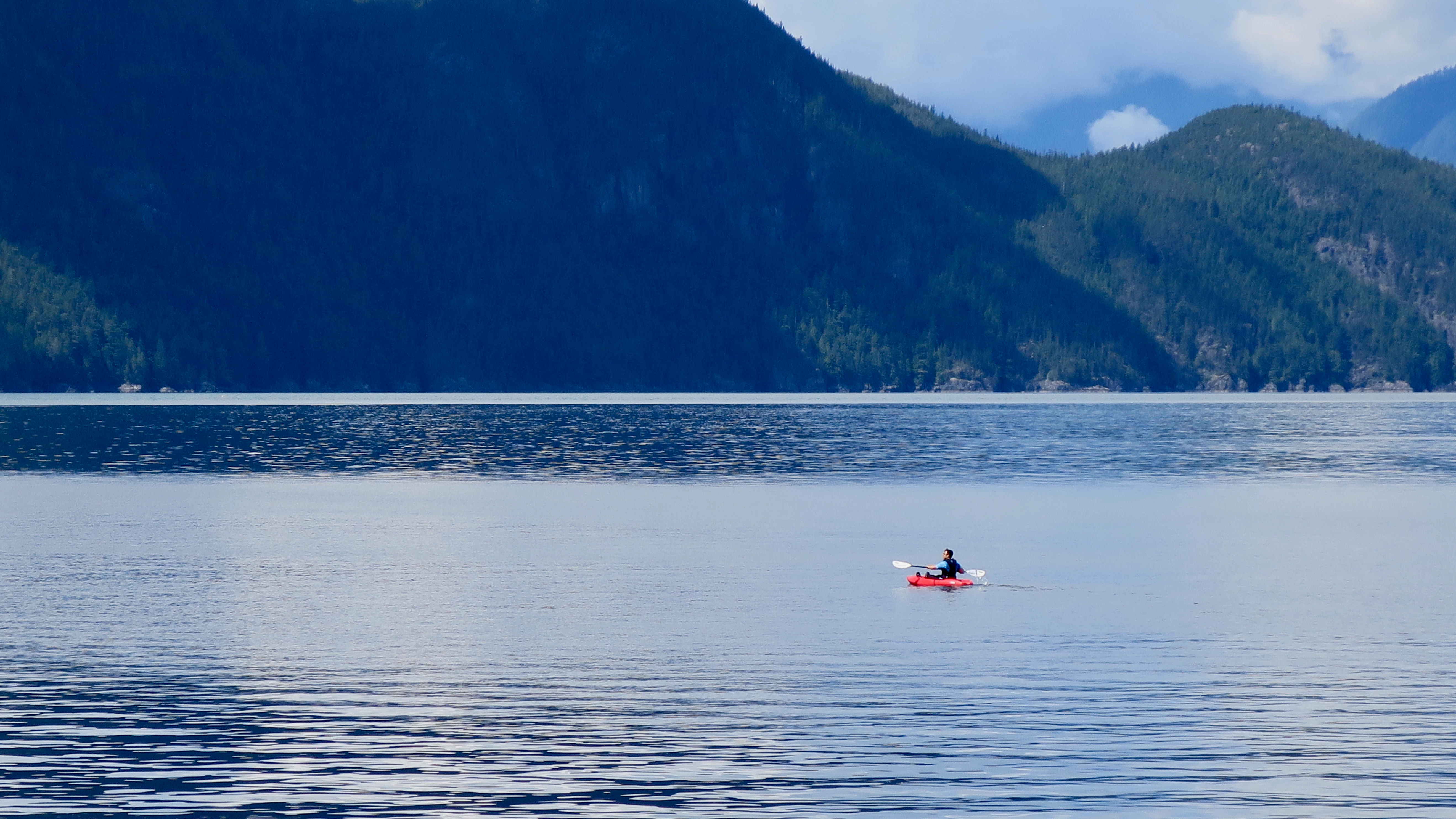 A wide shot of a calm body of water with a person in a small red kayak paddling in the middle; steep forest-covered mountains rise in the background.
