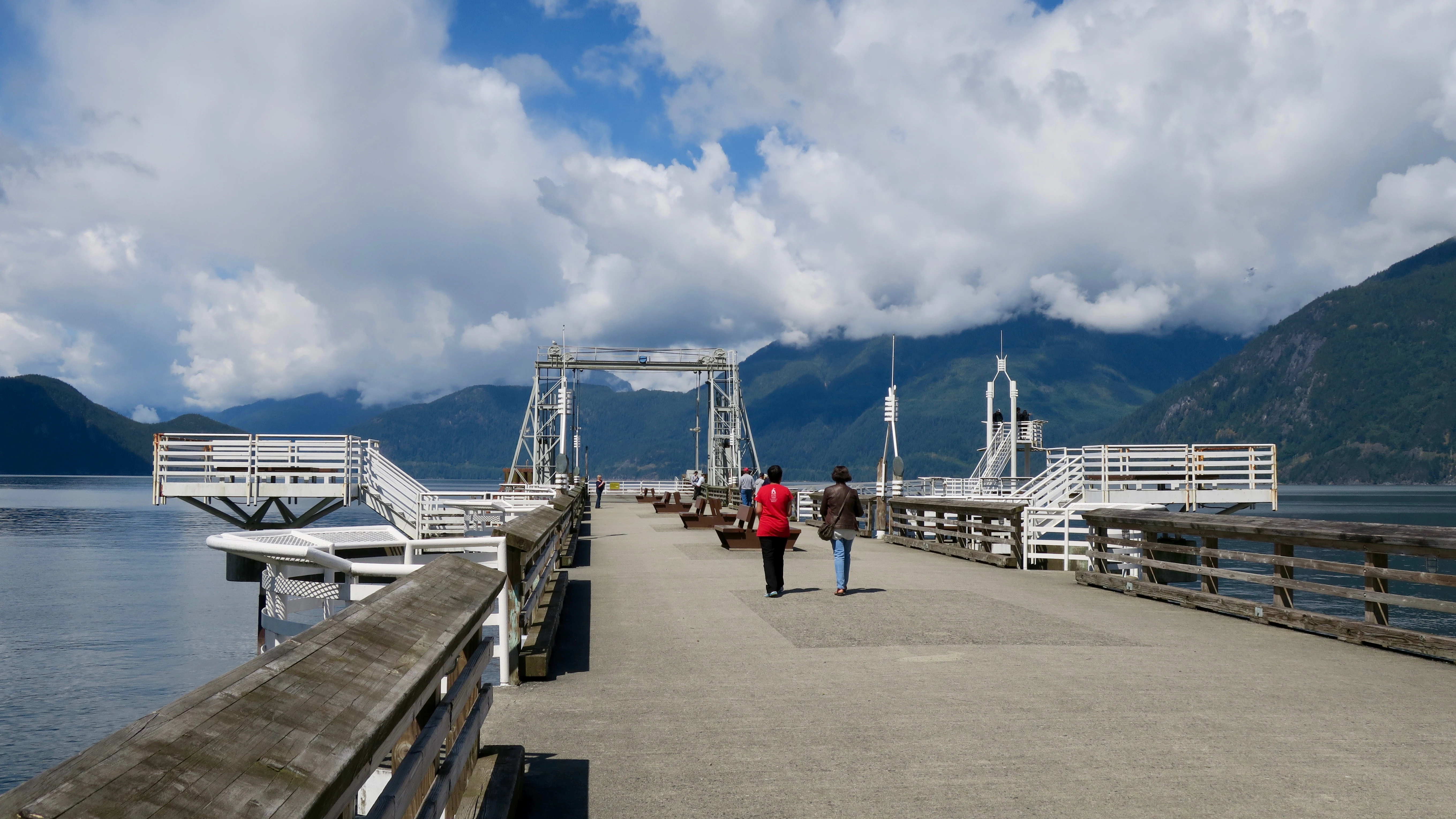 A wide shot from the perspective of a person walking on a long concrete pier, with wooden railings on the sides, extending into a calm body of water surrounded by mountains under a cloudy sky.
