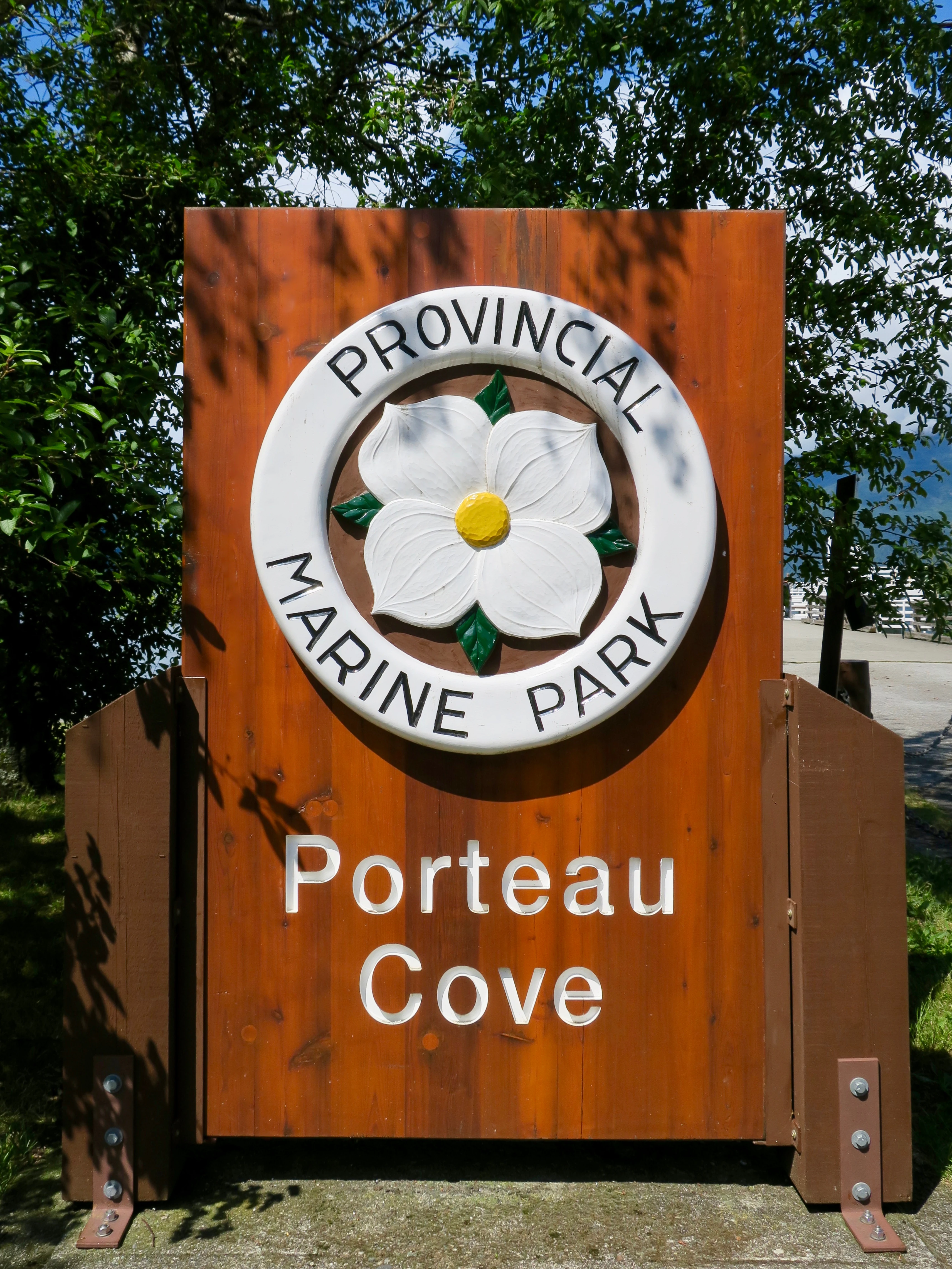 A wooden entrance sign for Porteau Cove Provincial Park features a circular emblem with a Pacific dogwood flower in the center, set against a background of green trees.