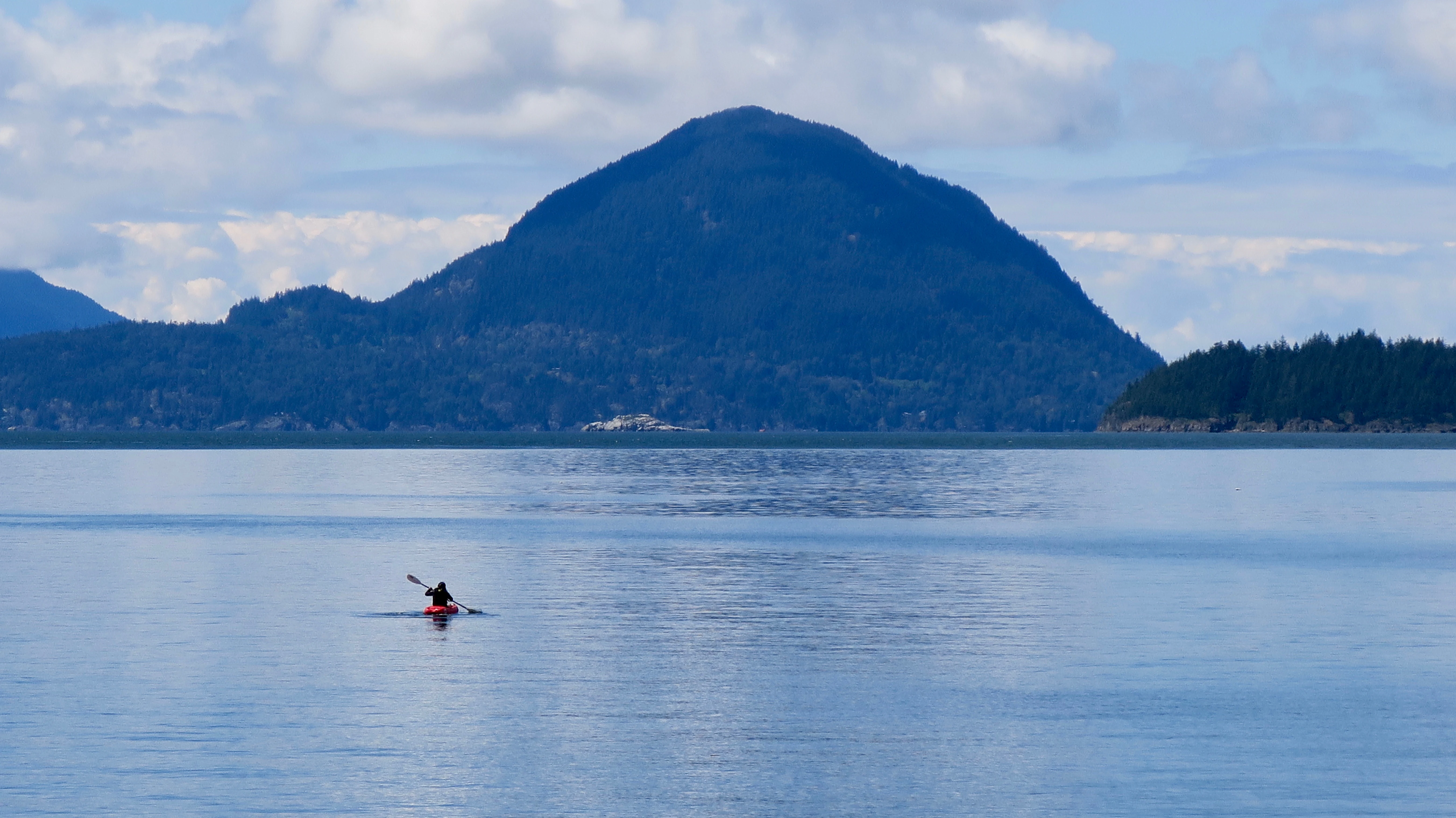 A wide, eye-level shot of a person in a red kayak on calm, blue water, with large, forested mountain peaks rising in the background under a cloudy sky.