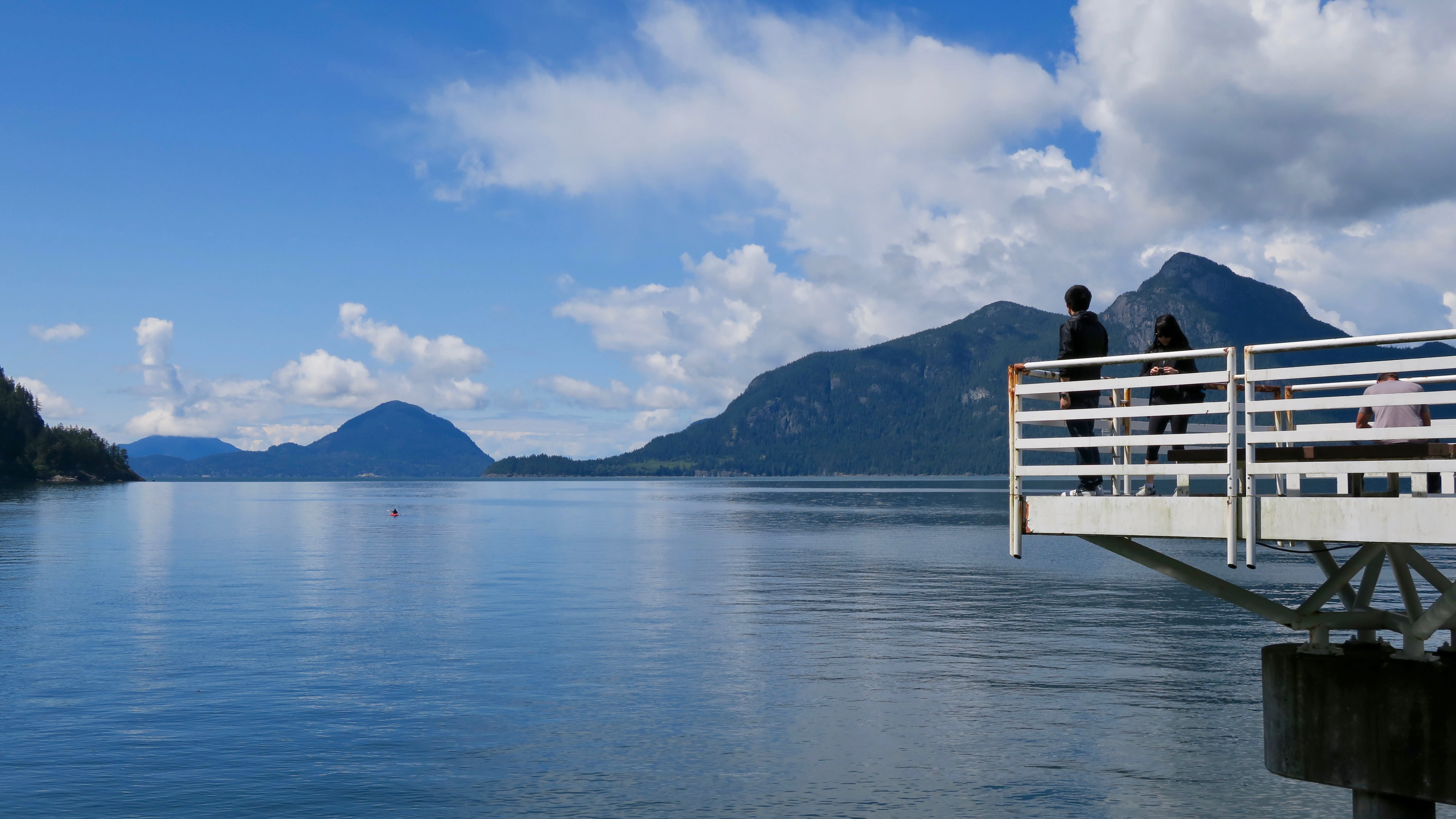 A wide shot of a calm blue sound with distant mountains under a partly cloudy sky, viewed from a white metal platform where two people stand.