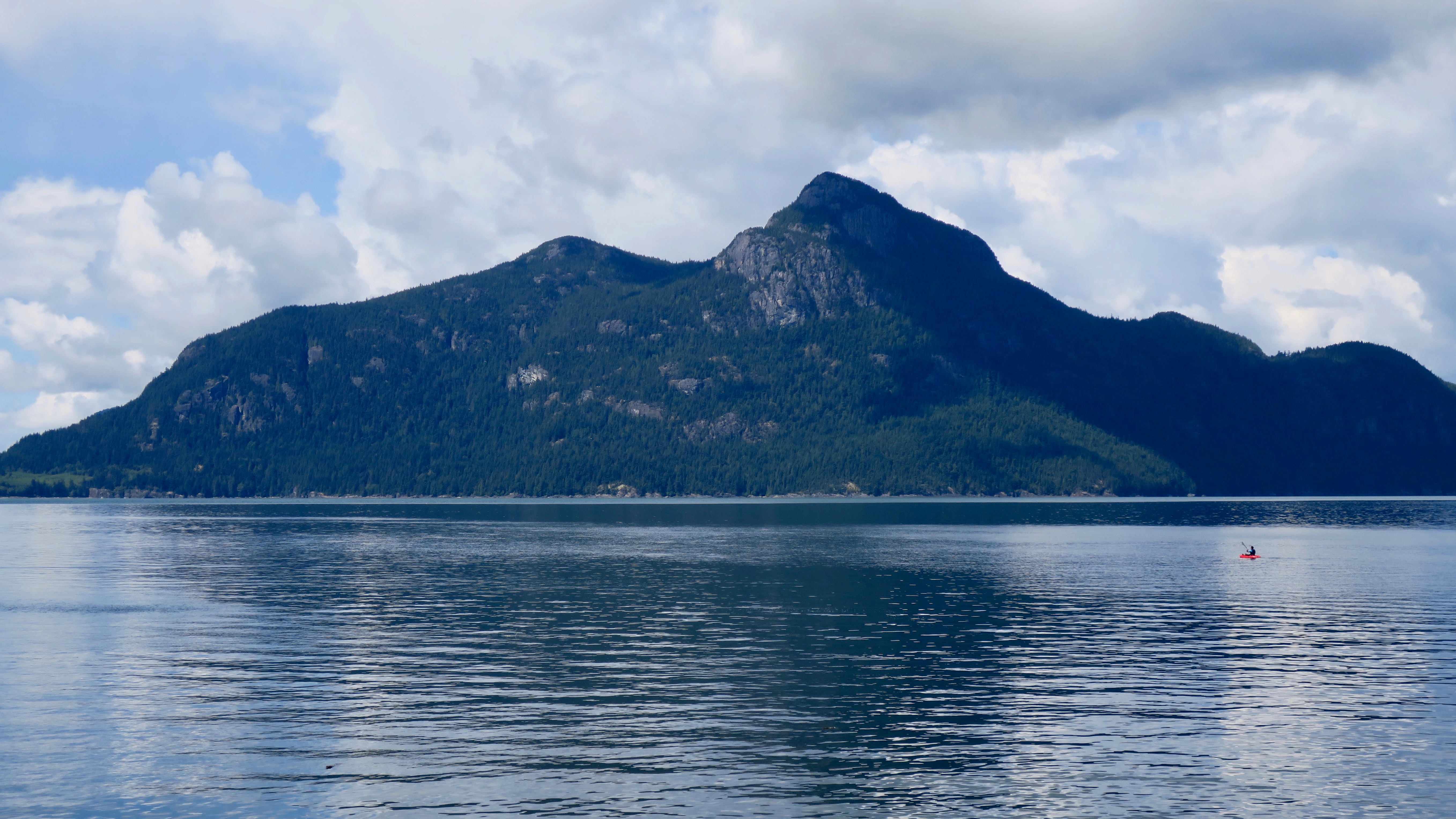 A calm, wide view of a large mountain rising directly out of the deep blue waters of a sound under a cloudy sky.