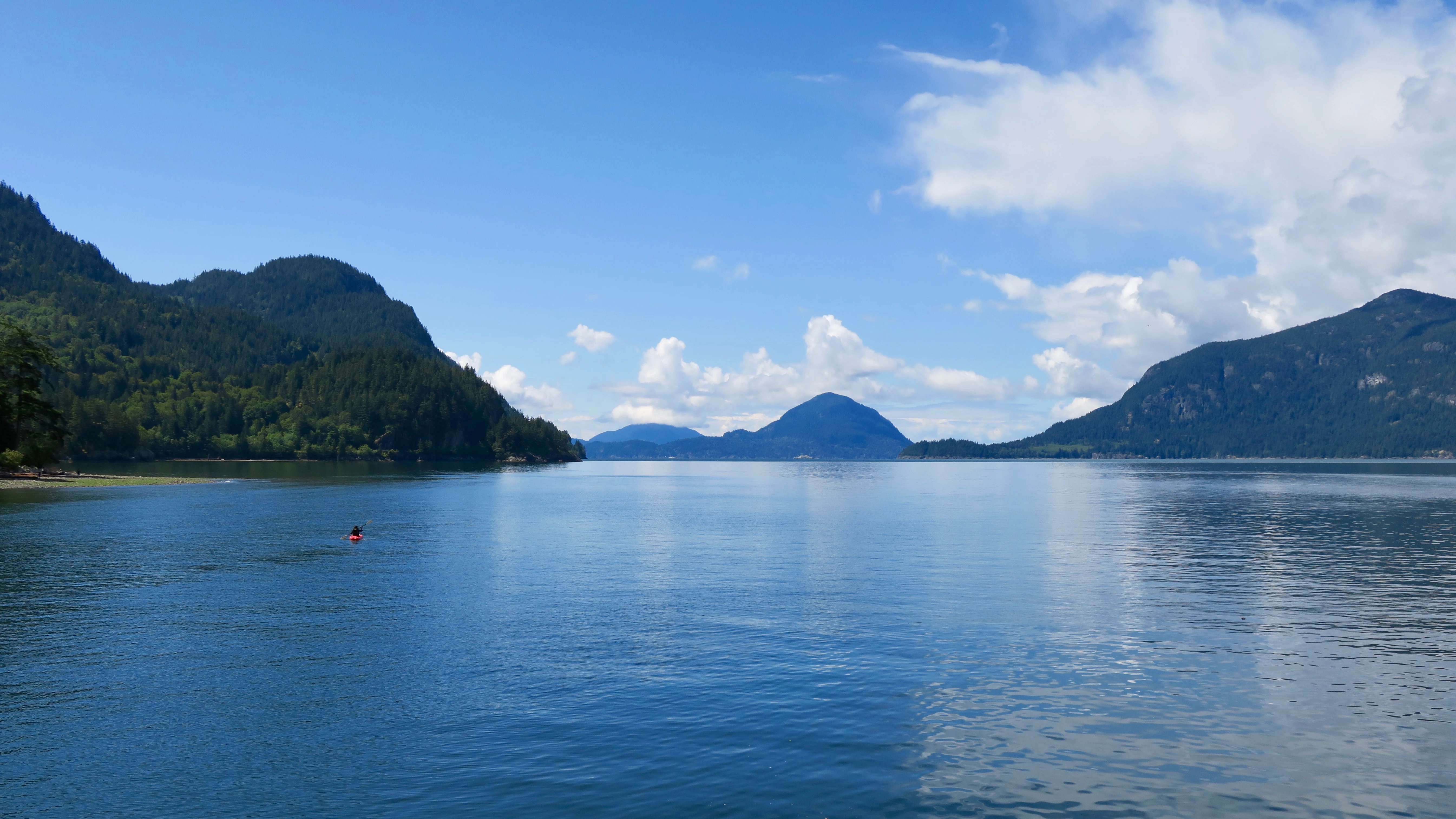 A wide shot looking across the calm, blue waters of Howe Sound toward distant forested mountains under a bright sky with scattered white clouds.