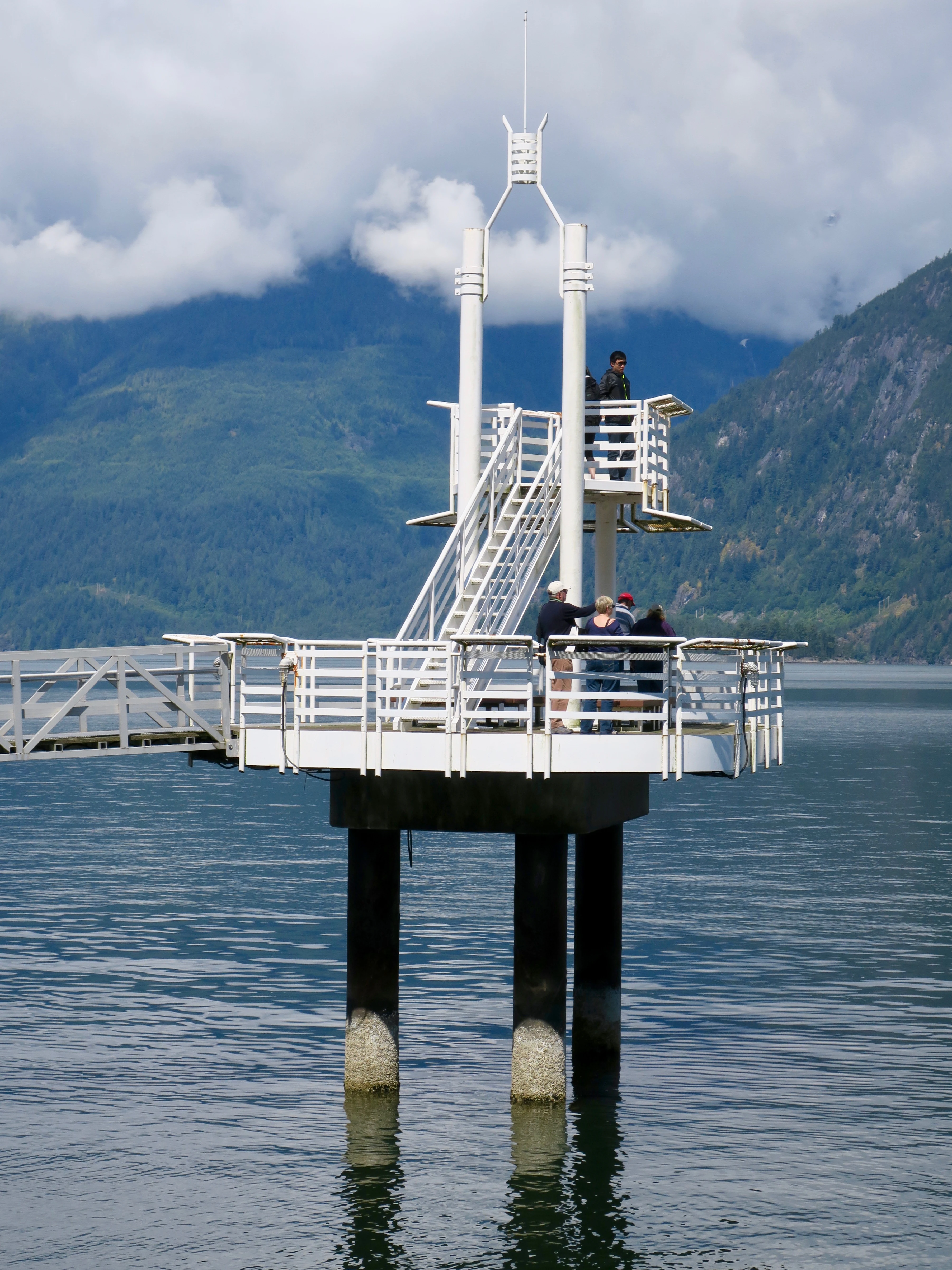A white metal observation tower extending over the water at Porteau Cove Provincial Park, with people standing on the deck and mountains in the background.
