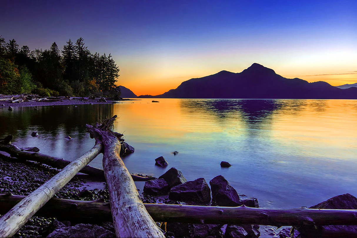 A peaceful landscape view across a calm inlet at sunset, with silhouetted mountains in the background and driftwood on a rocky shoreline in the foreground.