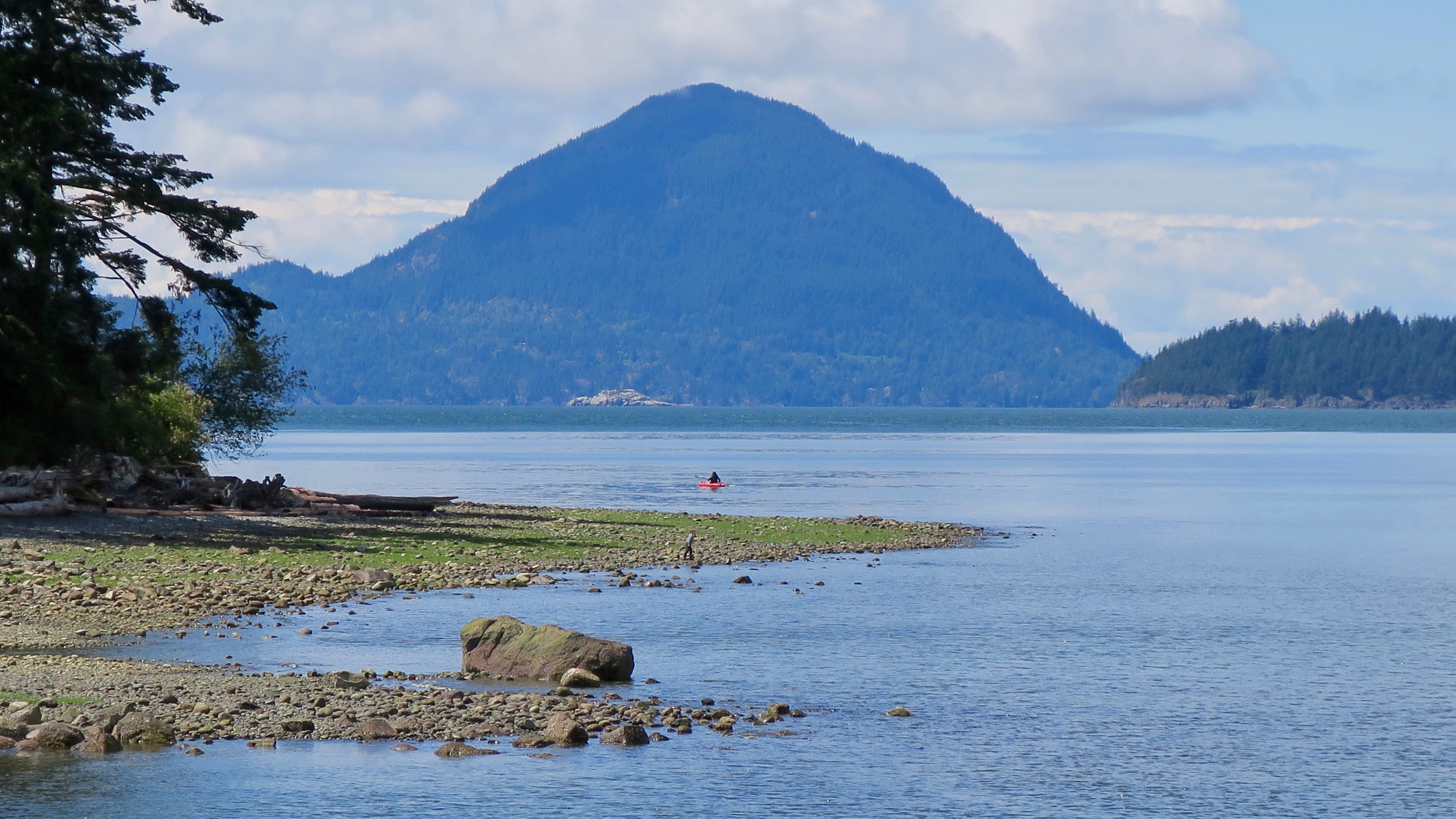 A peaceful coastal scene featuring a rocky shoreline, calm blue water, and a prominent mountain peak in the distance under a partly cloudy sky.