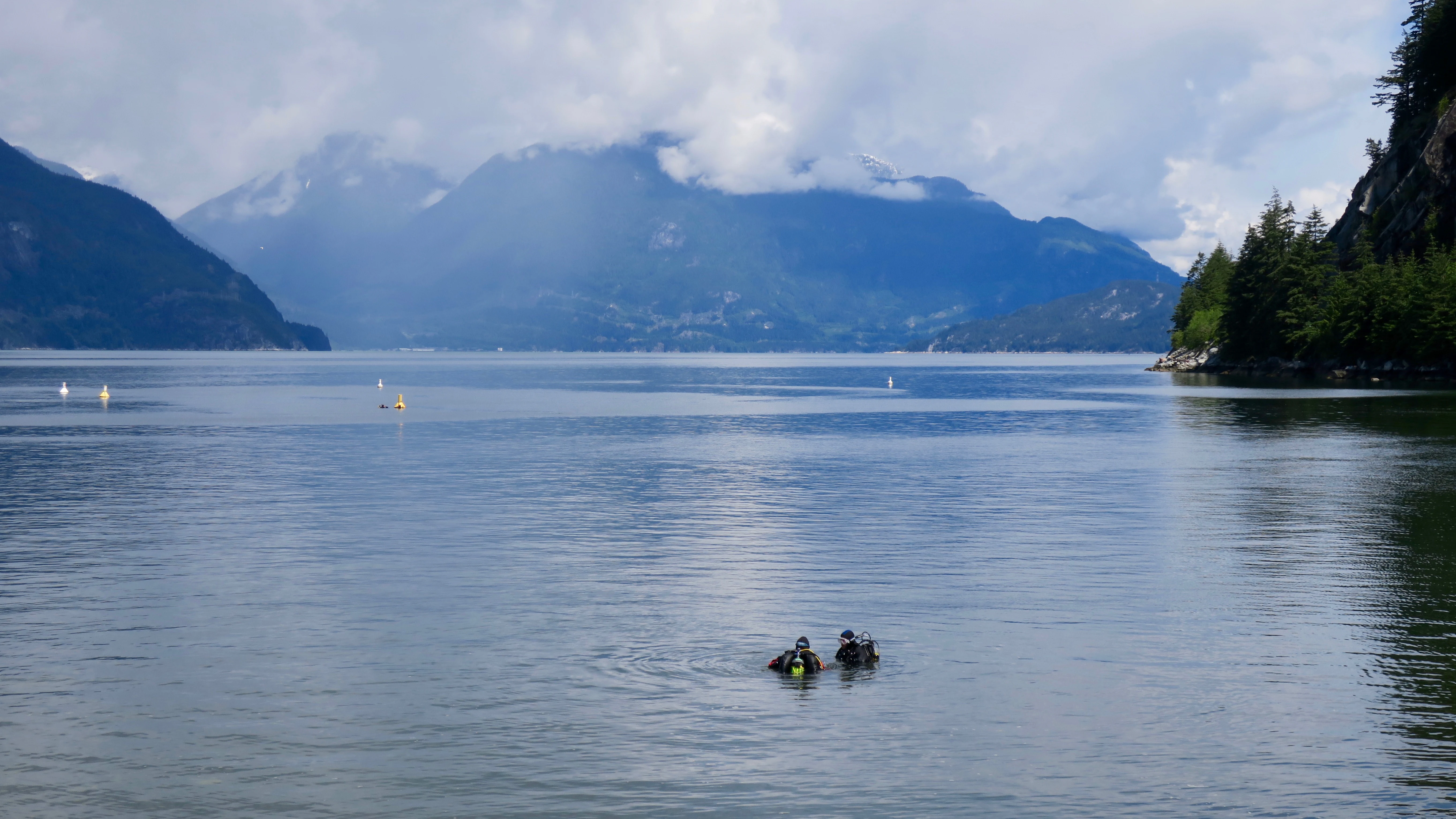Two scuba divers swim in the calm water of a bay, with dramatic mountains and clouds in the distance.