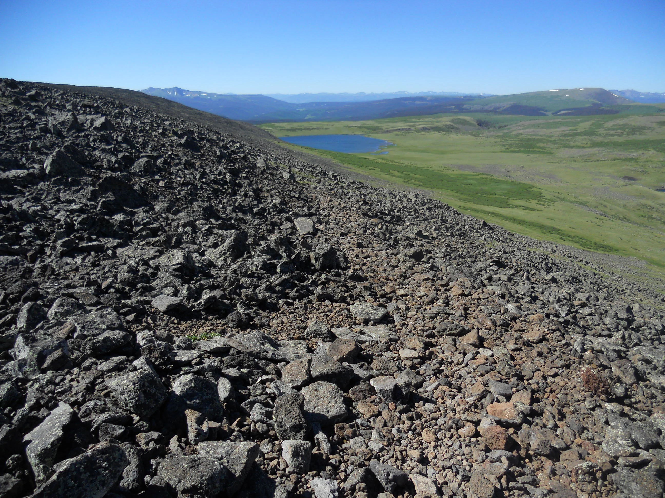 A wide, high-angle landscape shot of a blue alpine lake nestled in a green valley, with a large, rocky, gray volcanic scree slope in the foreground.