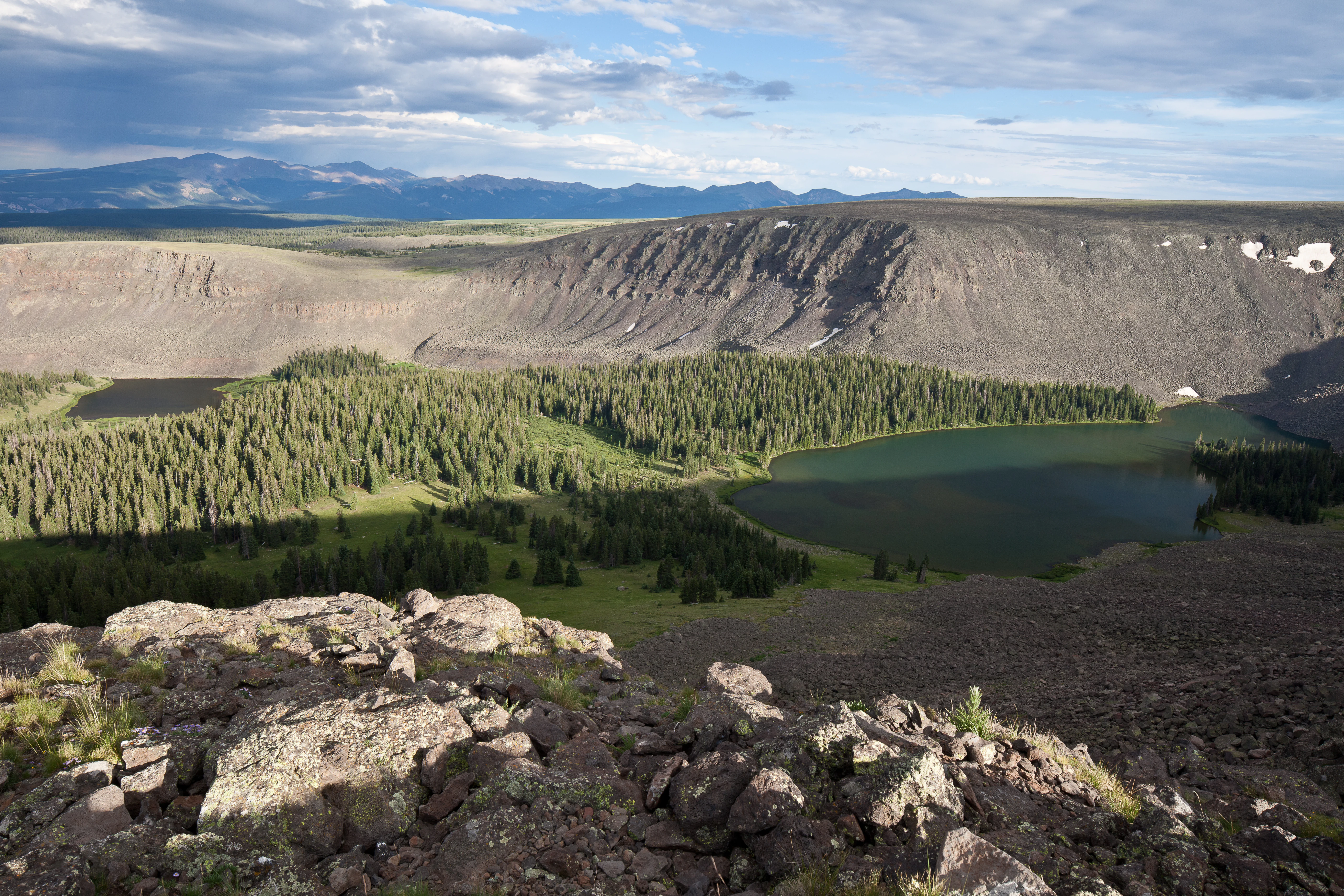 A high-angle view looking down into a valley containing a dark alpine lake surrounded by dense evergreen forests and steep volcanic cliffs.