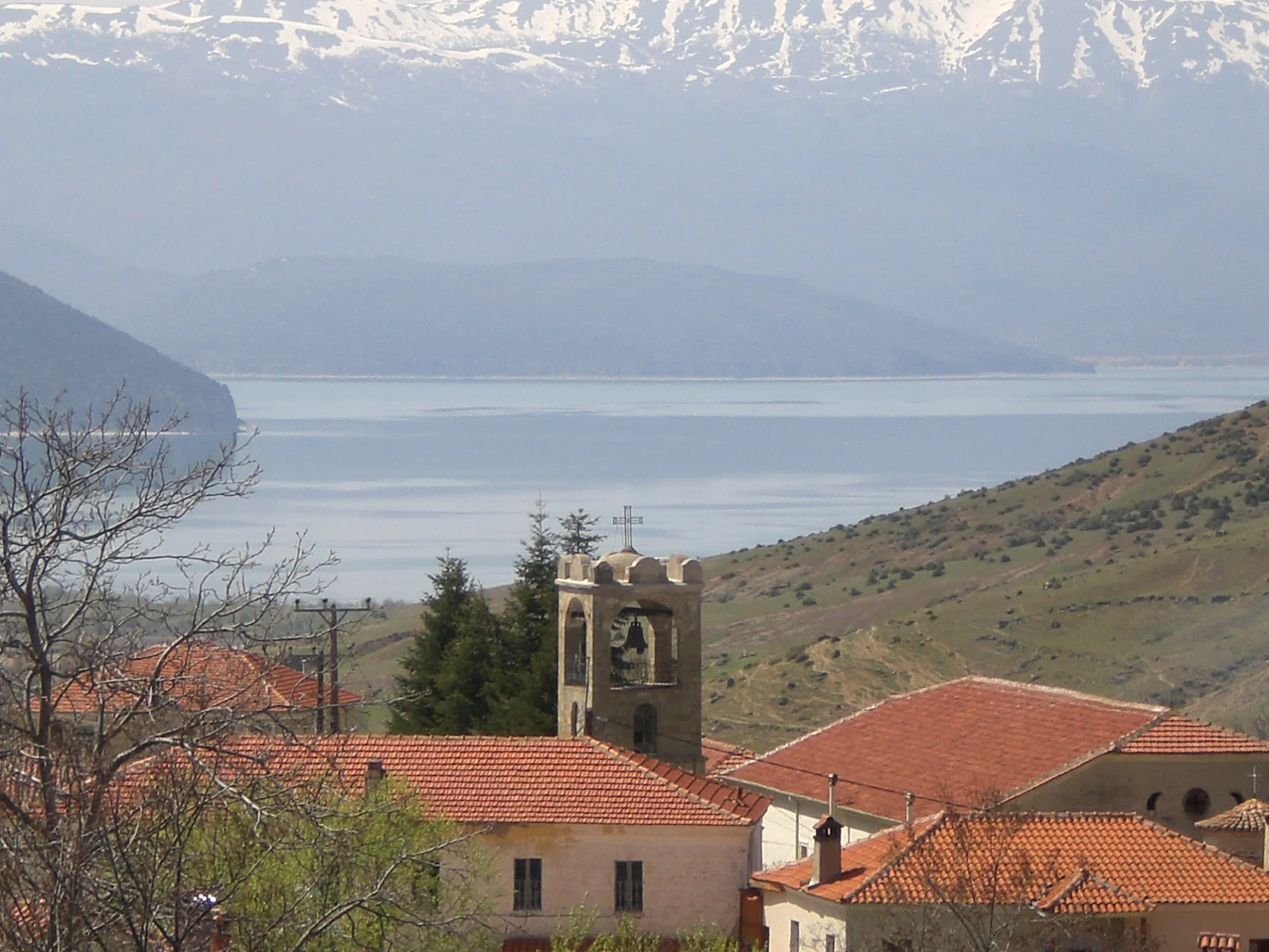 A view of the stone bell tower and terracotta roofs of Agios Germanos village in the foreground; with the waters of Lake Prespa and snow-capped mountains in the background.