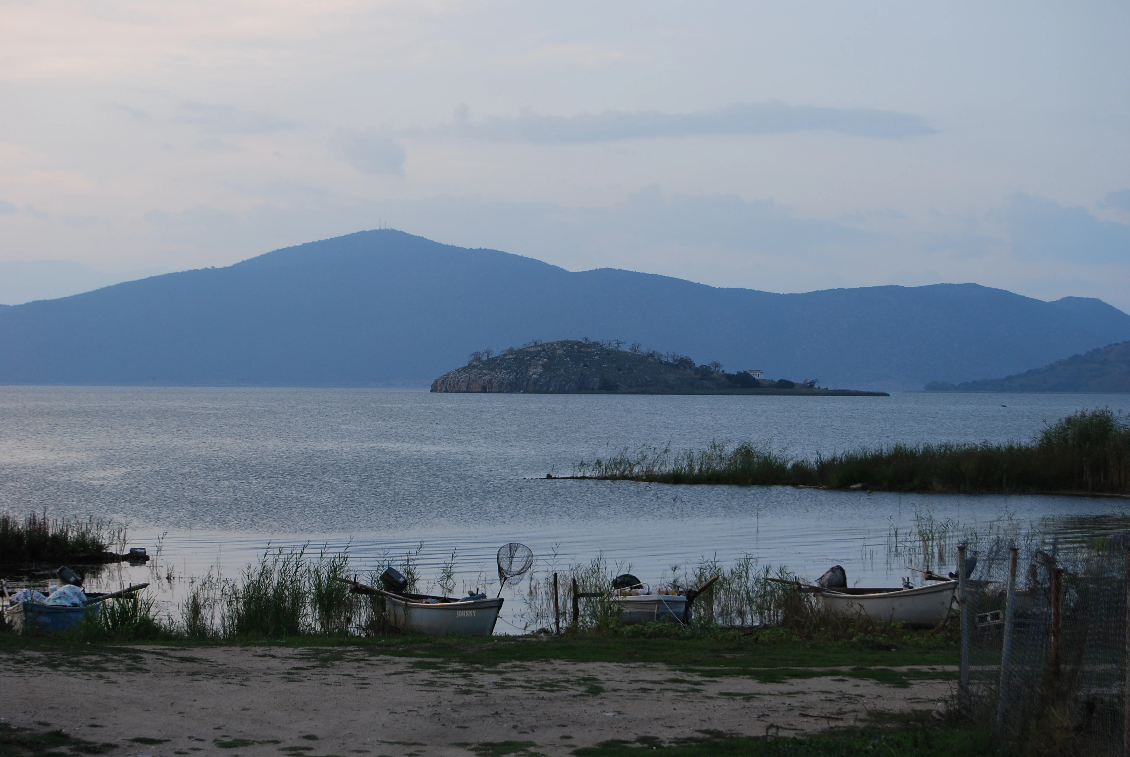 A calm lake landscape at dusk featuring Saint Achillios island in the middle distance, small fishing boats moored in the reeds in the foreground, and mountain silhouettes against a pale sky.