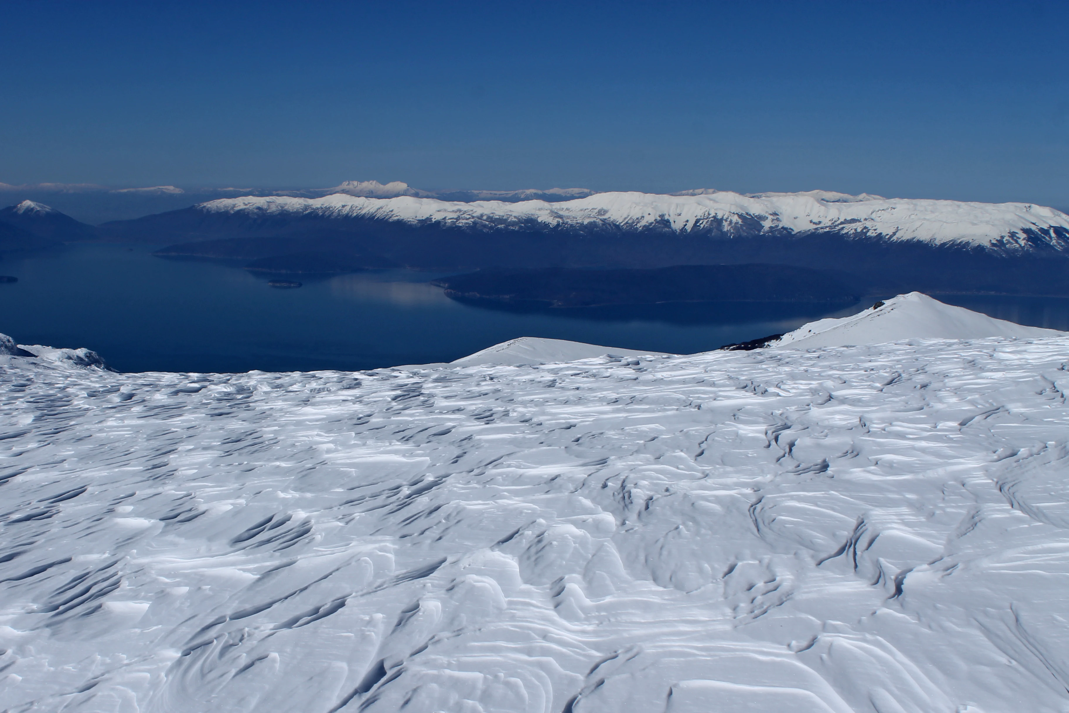 Snow-covered landscape with visible patterns, large lake, and mountain range under clear blue sky