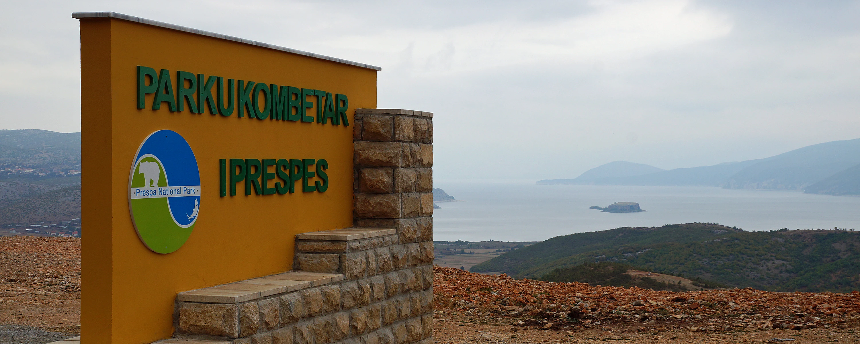 Yellow sign for Prespa National Park with Albanian text and logo, stone structure, and Lake Prespa visible in background