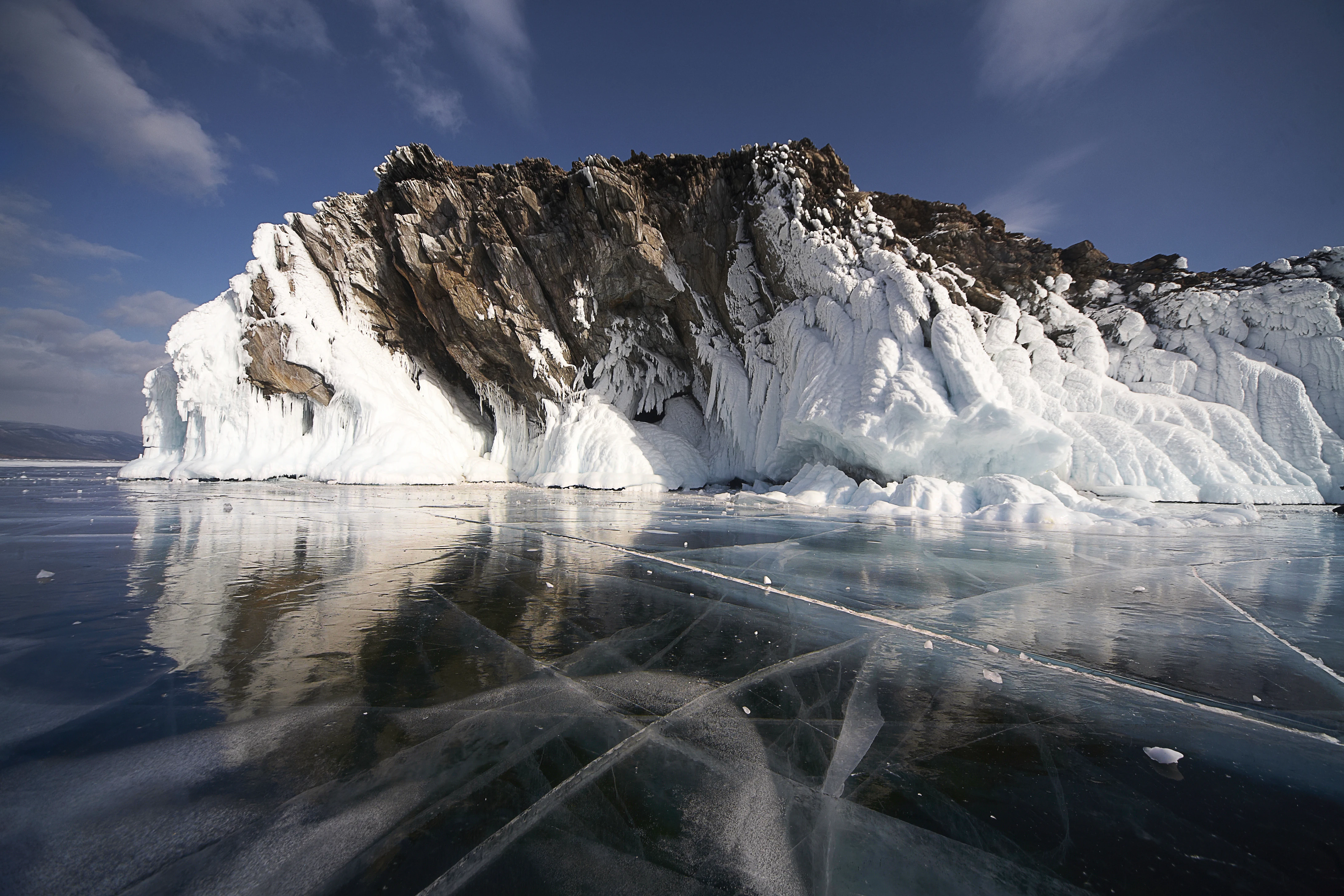 A jagged, rocky island cliff partially covered in ice, set against a deep blue sky and surrounded by the thick, clear, cracked ice of Lake Baikal in winter.