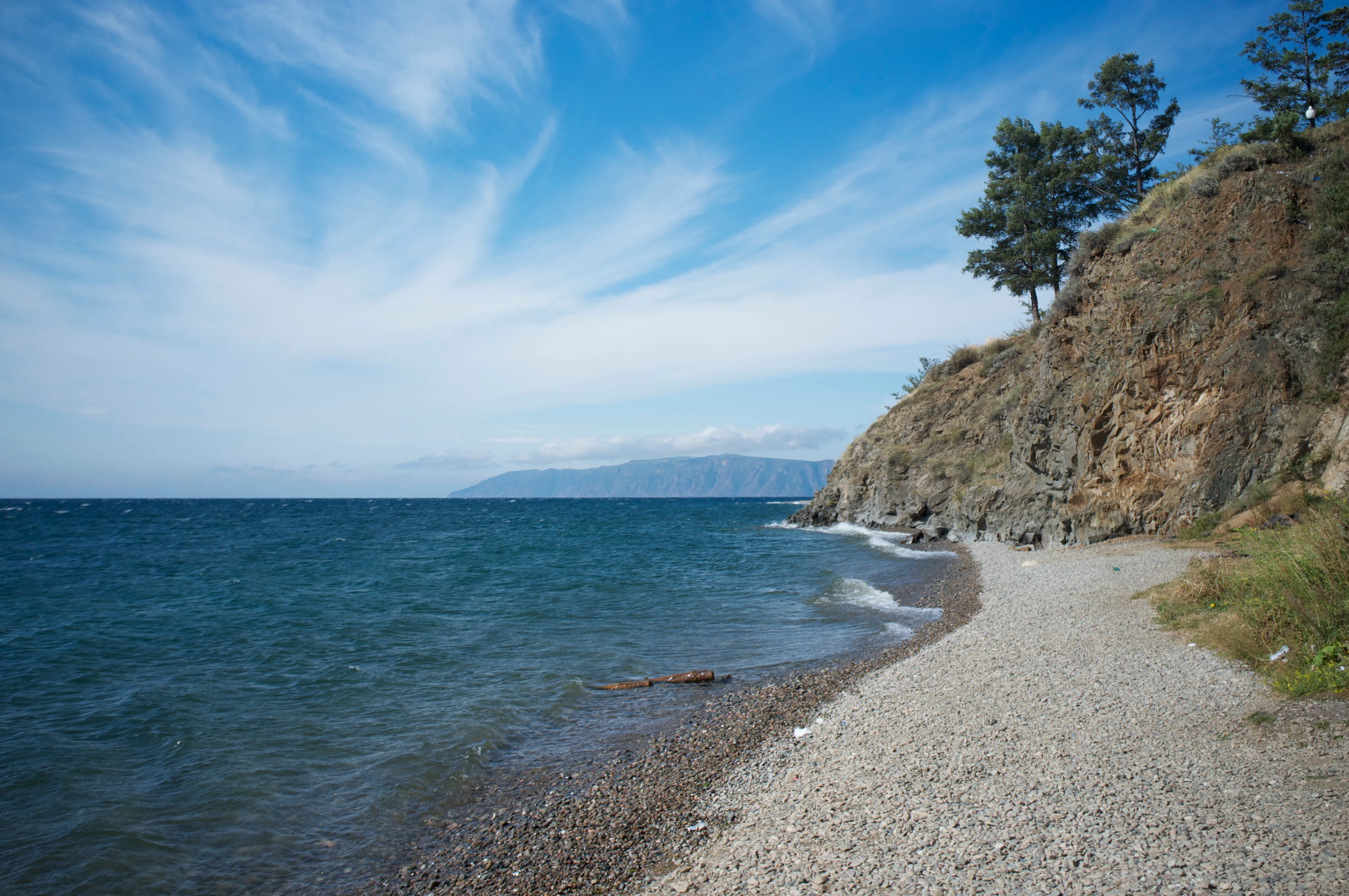 A wide angle view of a pebbled beach along Lake Baikal, with steep rocky cliffs topped with pine trees on the right and distant mountains across the lake under a blue sky.