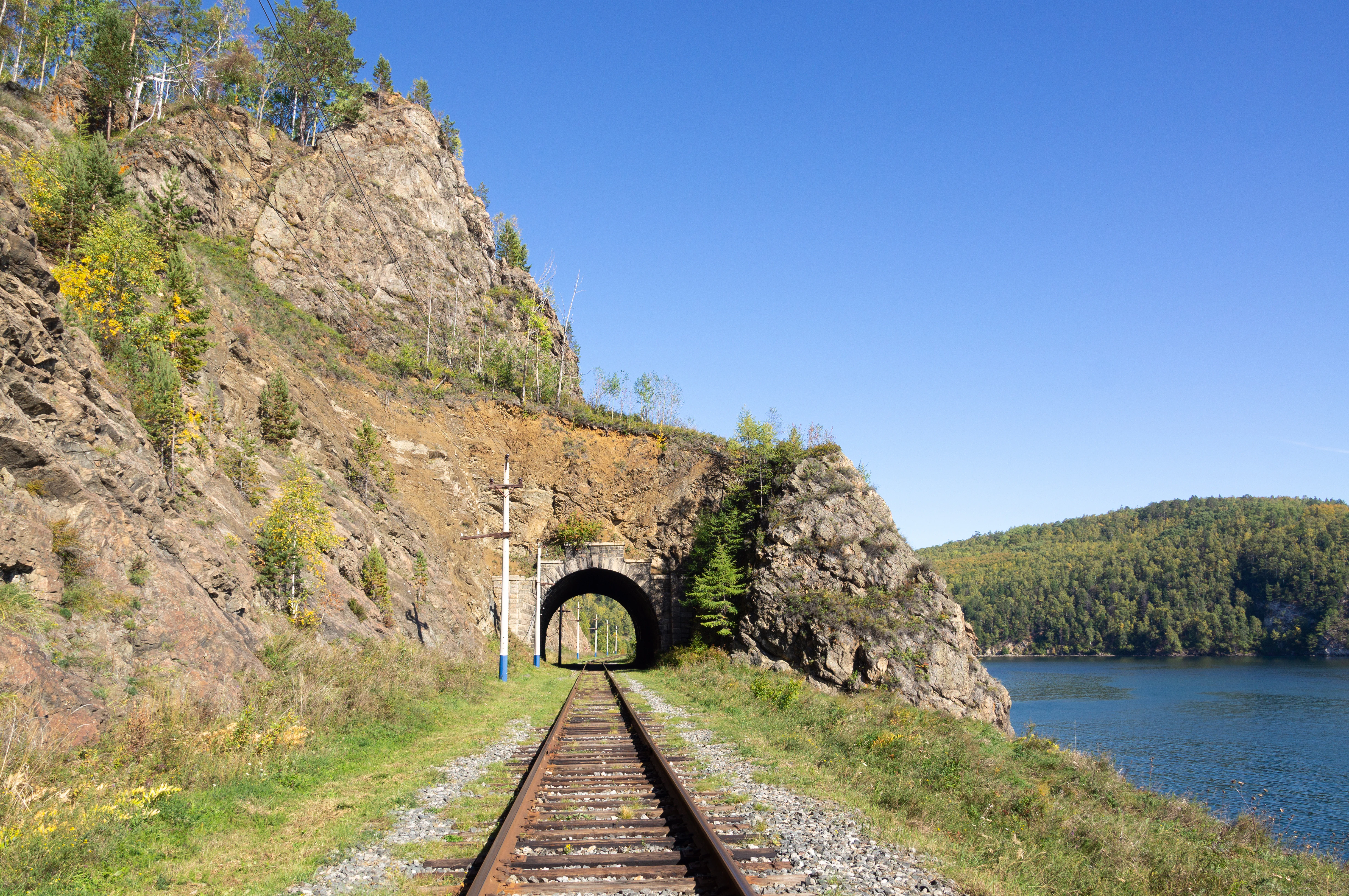 A railway track leads into a tunnel carved directly through a rocky cliffside, with the deep blue water of Lake Baikal visible to the right under a clear sky.