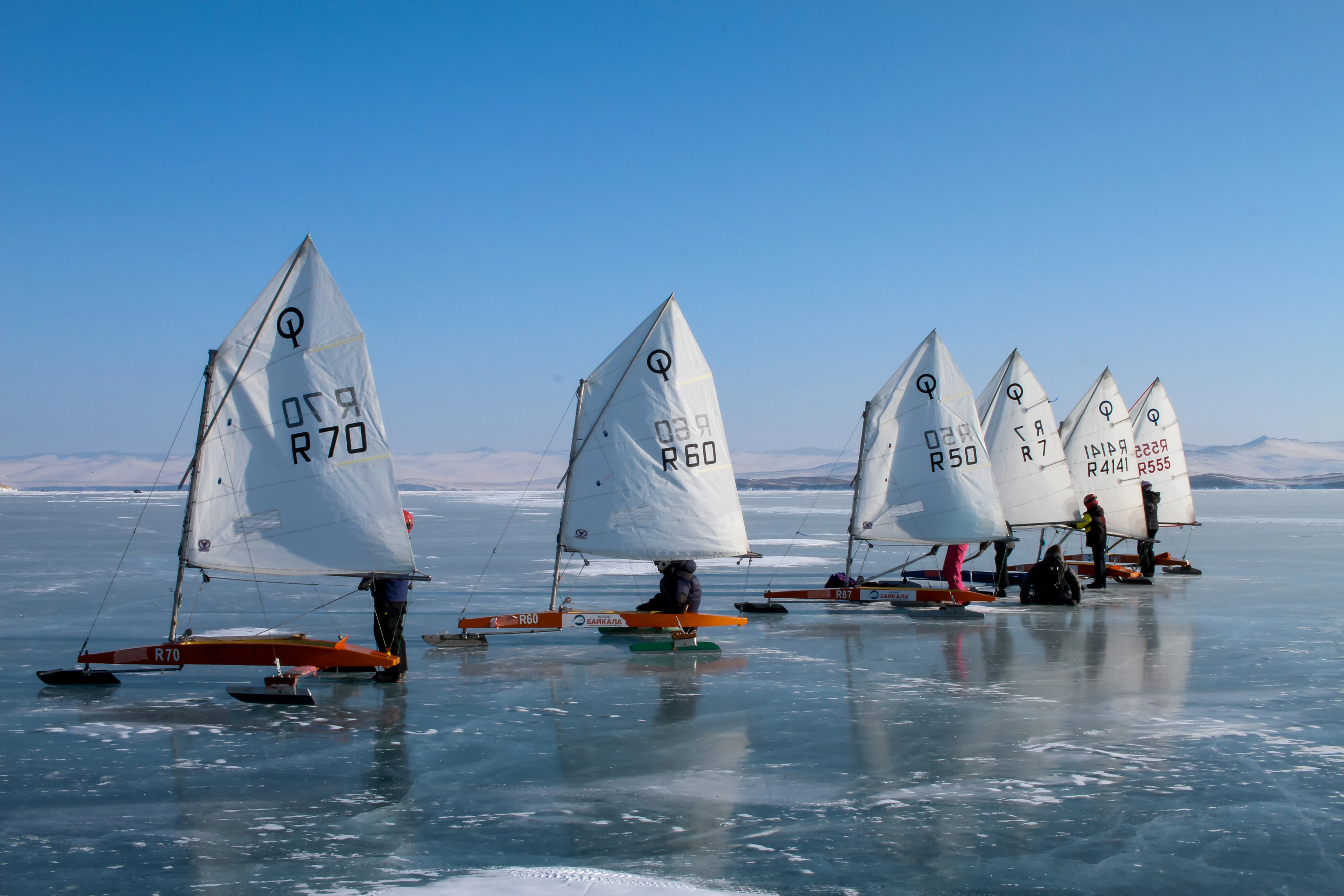 A group of ice boats with white sails lined up on the frozen, smooth surface of Lake Baikal on a clear winter day.