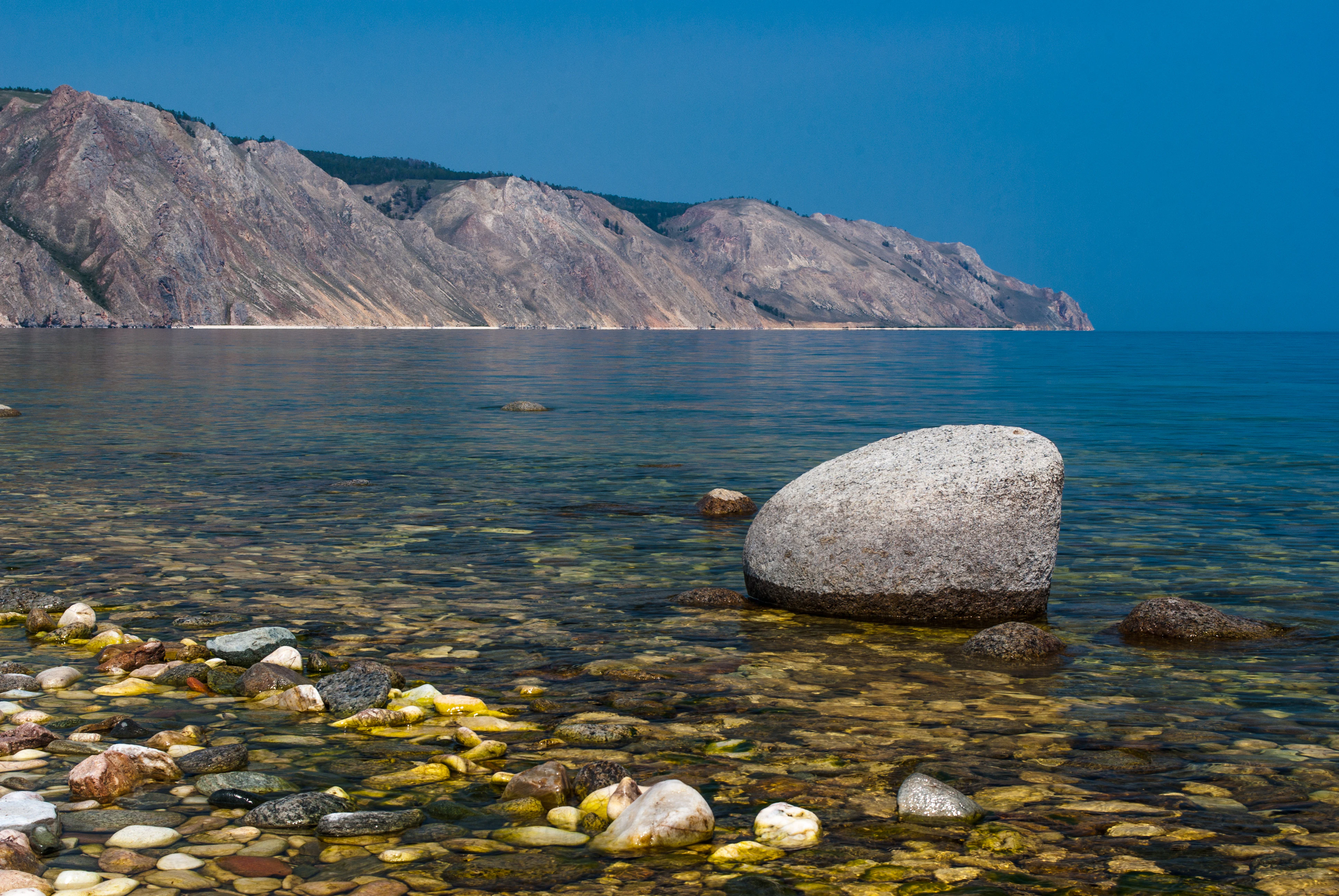 A clear view of a pebbled beach in the foreground with a large rounded boulder in the shallow water, leading to a calm blue lake and steep, rocky mountain slopes in the background under a clear sky.