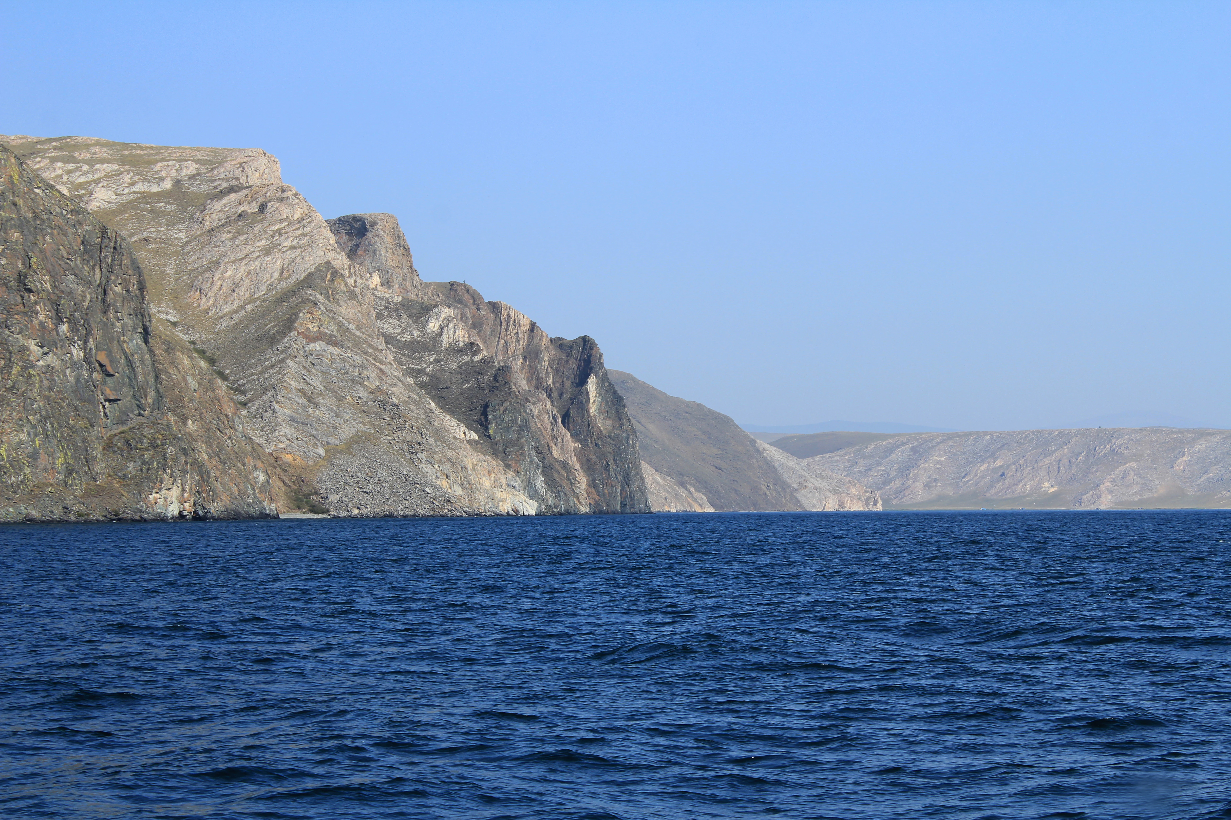 A wide view of rocky, layered cliffs meeting the blue water of Lake Baikal under a clear sky.