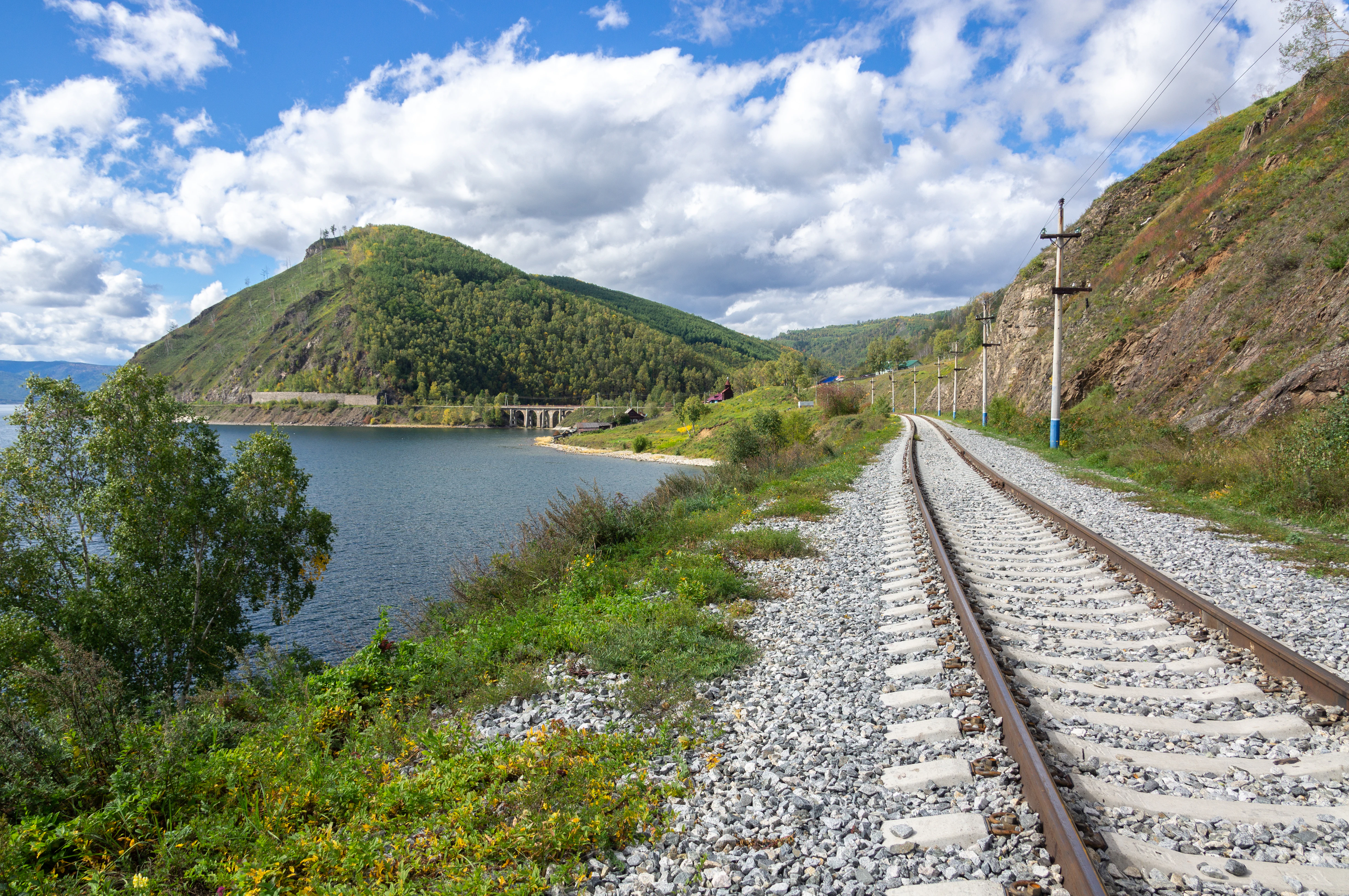 A railway track with gravel ballast runs along the shoreline of a large blue lake, with green hills and a forested mountainside in the background under a cloudy blue sky.